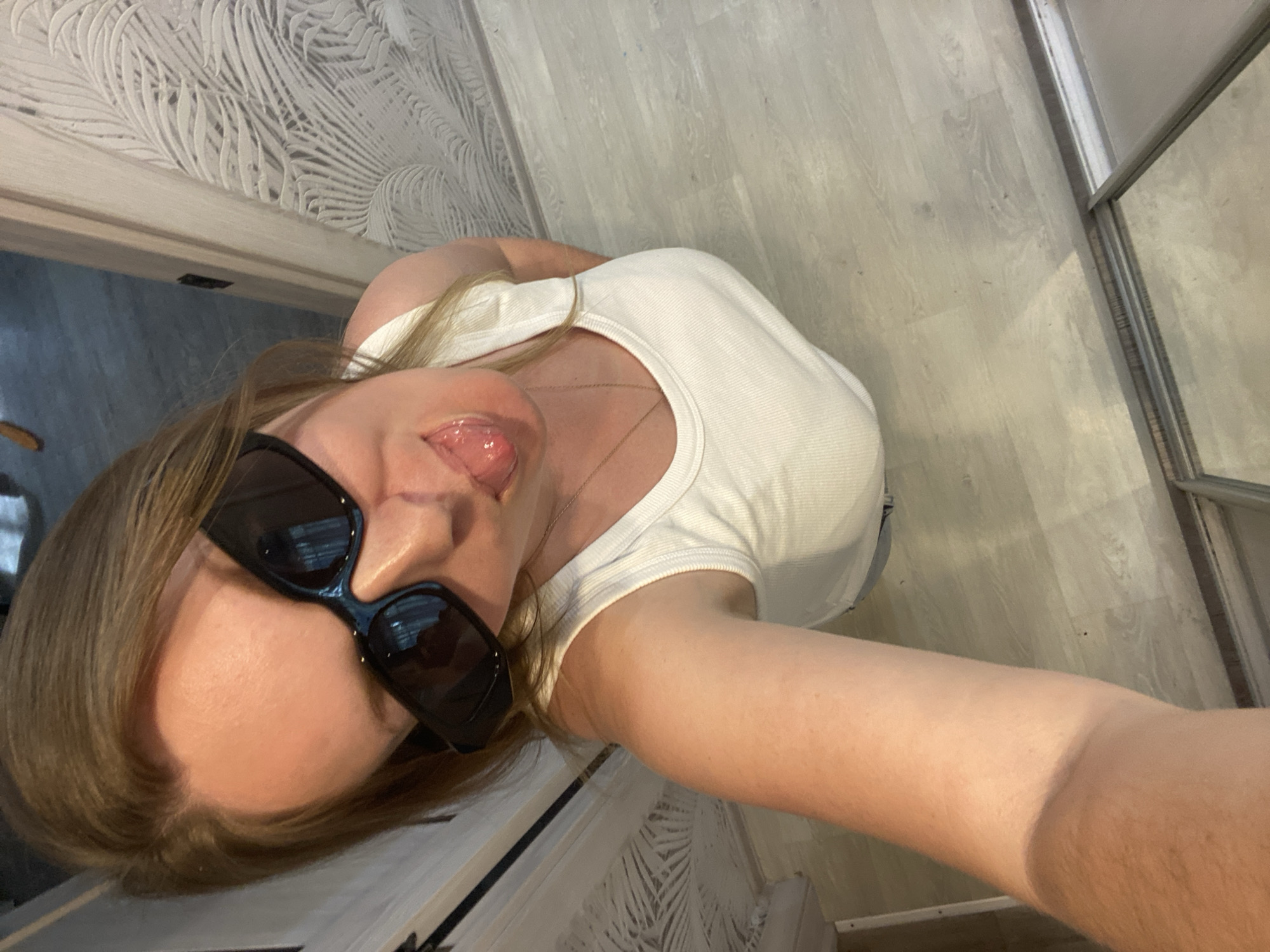 A woman wearing sunglasses and a white tank top is taking a selfie indoors. Her tongue is sticking out slightly. The background includes a patterned wall, a wooden floor, and a closet or wardrobe with sliding doors. The location appears to be inside a building, possibly an apartment or house. There is no information about the city of Noviy, Russia in the image.