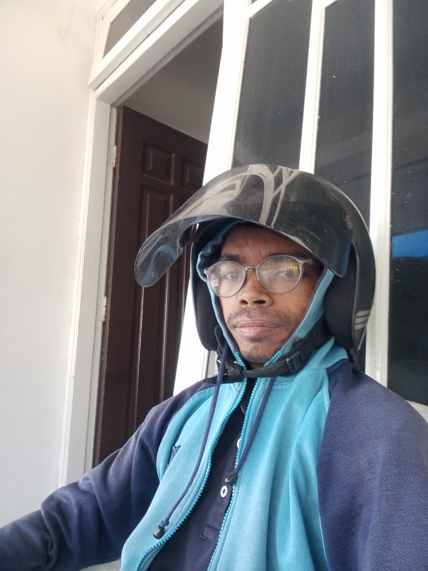 In Surabaya, Indonesia, a person wearing a dark helmet and a light blue and dark blue hooded jacket is seen in a close-up shot. The subject has on round glasses, and a dark brown door is visible in the background. The scene appears to be outdoors, possibly near a building entrance.