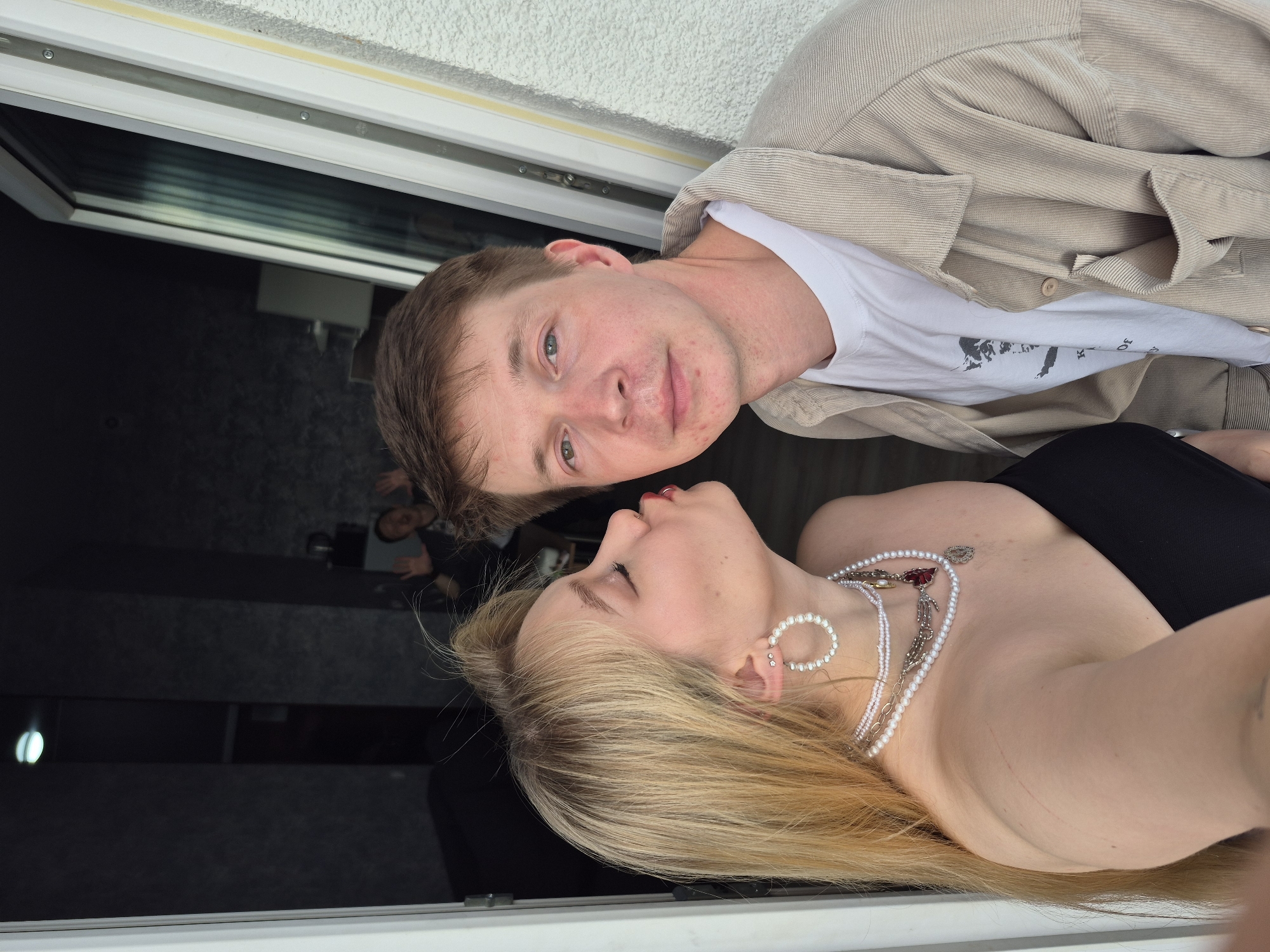 A medium shot captures a young woman with blonde hair leaning in to kiss a young man. The woman is wearing a black strapless top and multiple pearl necklaces, as well as large hoop earrings. The man is wearing a beige corduroy jacket over a white t-shirt. They are standing in front of an open doorway, with the man facing the camera and the woman's face obscured as she kisses him. In the background, through the doorway, another person can be seen with their hands up. The exterior wall of a building is visible to the right. The scene appears to be in an urban setting, possibly a residential building in Izhevsk, Russia, given the context of the location.