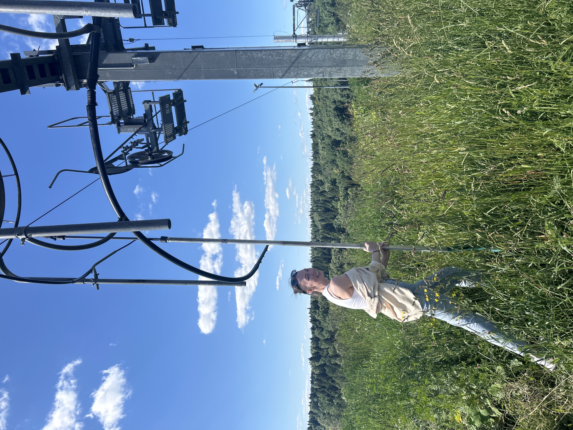A young woman is shown standing in a grassy field next to a ski lift structure. She is wearing a white tank top, a light-colored open-front shirt, blue jeans, and sunglasses pushed up on her head. She is holding onto a pole that extends from the ski lift structure. Her body is angled towards the viewer, and she is smiling, appearing happy.
The scene is set in a lush, green field with tall grass and some small yellow flowers. In the background, a dense forest stretches across the horizon under a bright blue sky dotted with fluffy white clouds. The ski lift structure, made of metal, is prominent on the left side of the image. It is composed of various poles, cables, and mechanical parts, including a pulley wheel.
The lighting suggests it is daytime, likely late morning or afternoon, with the sun casting clear shadows. The weather appears to be warm and sunny, conducive to outdoor activities. The presence of the ski lift suggests a location that might be a ski resort or a mountainous area that is utilized for skiing during colder months. Given the location context of Krasnogorsk, Russia, this scene could be depicting a summer day at a recreational area. There is no visible text in the image.