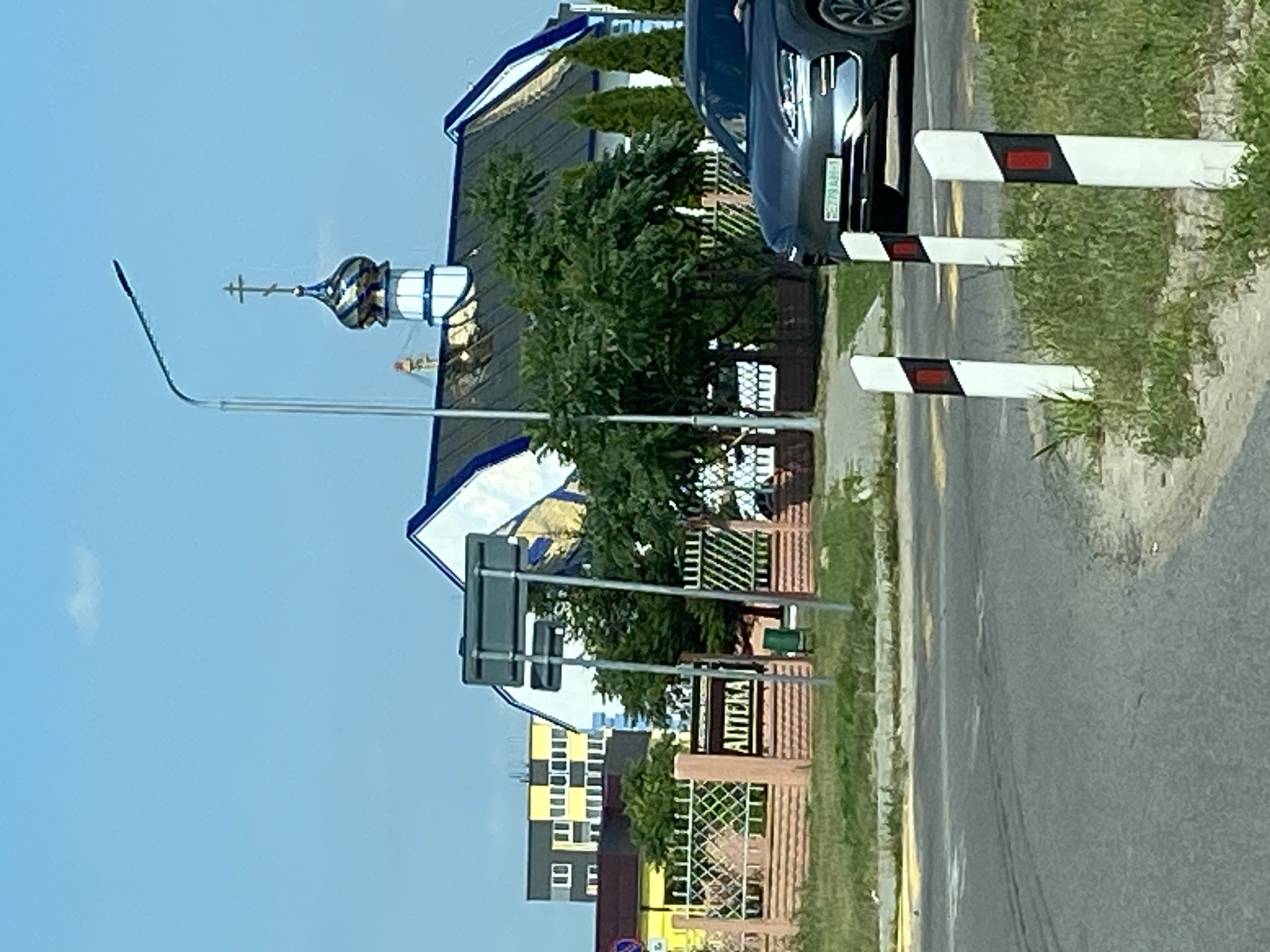 The image shows a view of a street scene with a building and a car. In the background, there is a white building with blue trim and a golden dome topped with a cross, which appears to be a church or religious building. The building has a dark roof. In front of this building, there is a fence with a sign that reads "АПTEKA," indicating a pharmacy. Behind the fence, there are trees and a modern building with yellow and grey panels. A dark-colored car is visible in the foreground, partially obscuring a row of white and red traffic markers. The sky is clear and blue with a small white cloud. The environment appears to be an urban or suburban setting in the vicinity of Матыкальскі сельскі Савет, Belarus.