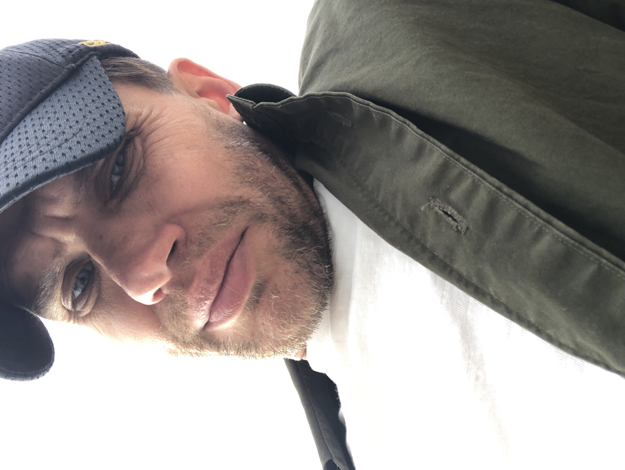The image shows a close-up, low-angle shot of a man wearing a dark baseball cap and a green jacket over a white t-shirt. He has a beard and is looking towards the camera with a serious expression. The background is a bright white, suggesting an outdoor setting with strong lighting, possibly against a clear sky or a white wall. There are no identifiable landmarks or environmental features to determine a specific location in Russia.