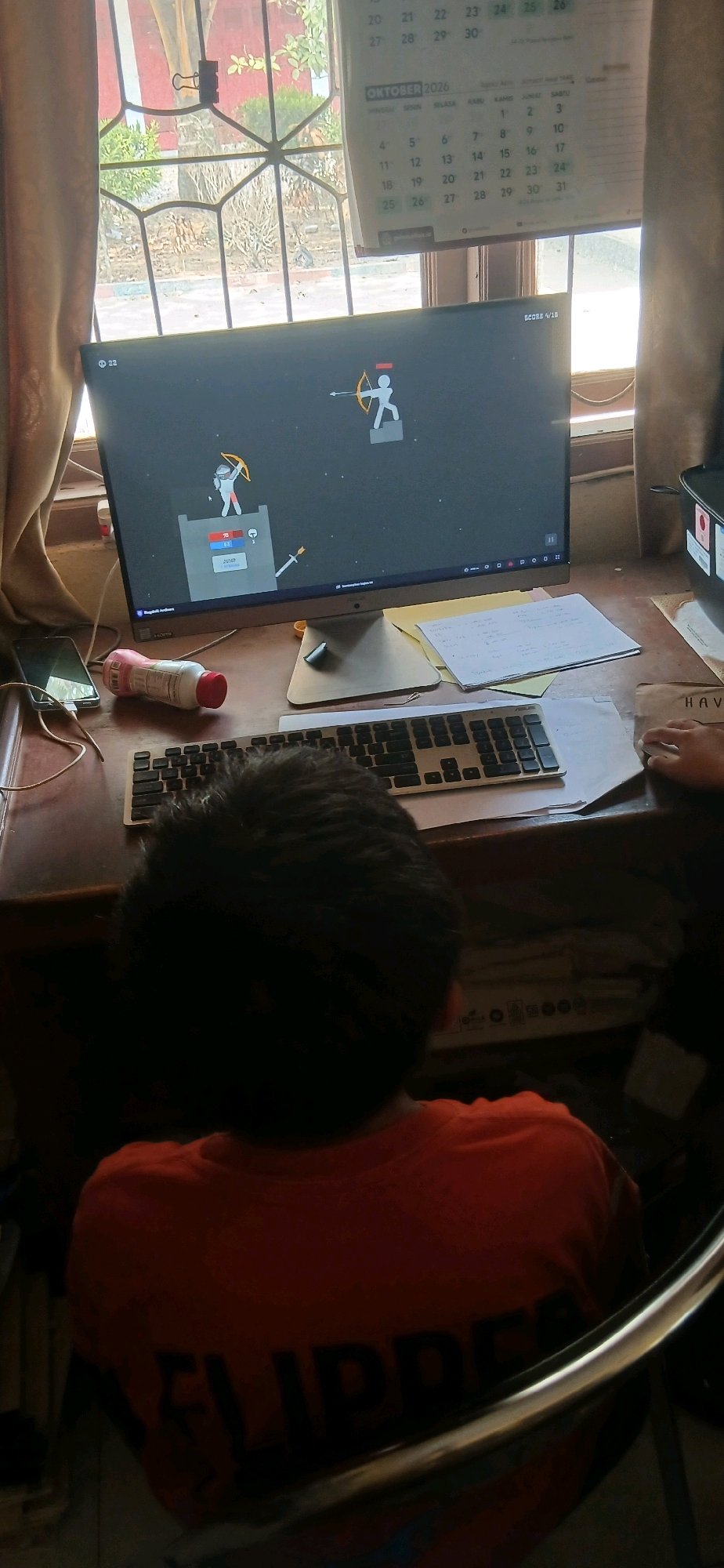 The image depicts a young person wearing an orange t-shirt sitting at a desk in front of a computer monitor. The computer screen displays a game with stick figures, one of whom is an archer. A keyboard is positioned in front of the monitor. On the desk, there is also a smartphone, a bottle, and some papers. In the background, a window with a view of trees and a calendar showing "OKTOBER 2026" are visible. The scene appears to be an indoor setting, likely a home or study area. The location is specified as Metro City, Indonesia.