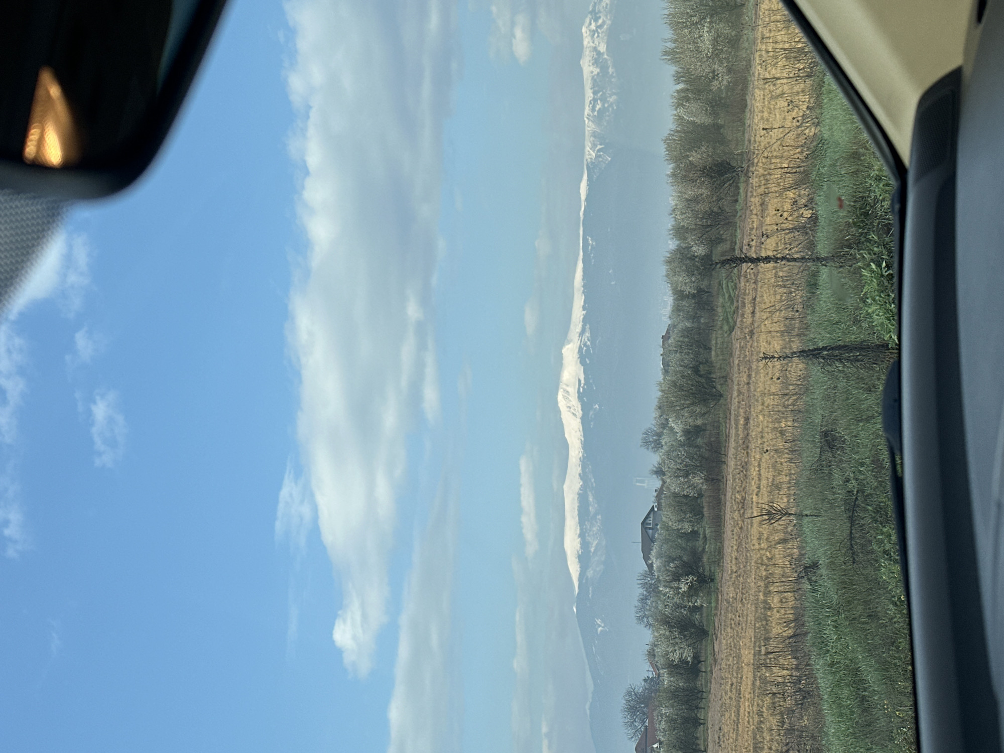 The image is a slightly tilted, horizontal view from inside a car. On the left, there is a rearview mirror with an illuminated part visible. The sky is blue with white clouds, and in the distance, there are snow-capped mountains. The right side of the image shows a field with trees and grass, partially visible through the car window. The location is not determinable from this image.