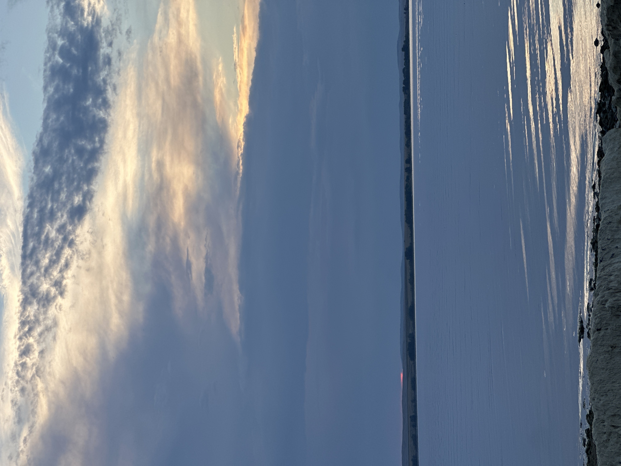 The image depicts a tranquil, wide-body of water, possibly a lake or reservoir, at twilight. The sky is a dramatic canvas of clouds, with streaks of warm, golden light breaking through darker, moody blues and grays. The water's surface is calm, reflecting the sky's colors and creating subtle patterns of light. A distant horizon line is marked by a low-lying landscape, sparsely dotted with trees and silhouetted hills. A thin sliver of bright red, indicative of the setting sun, is visible on the horizon. In the foreground, the edge of the body of water is visible, with rough, rocky terrain and some lighter-colored, possibly mineral-rich deposits. There are no people, animals, or man-made structures immediately apparent in the scene. The overall impression is one of natural beauty and serene, open space. The provided context identifies the location as Energetik, Russia.