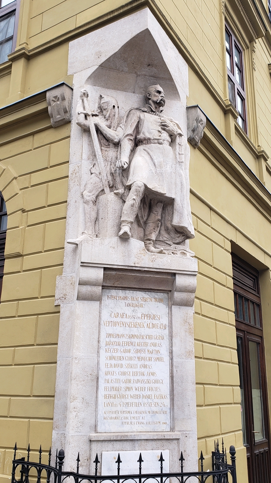 The image shows a relief sculpture on the exterior of a building. The sculpture depicts two figures, one appearing to be a soldier or warrior holding a sword and shield, and another figure in military attire with his hand on his chest. Below the sculpture is an inscription in Hungarian. The building is yellow with brown window frames and a dark brown wooden door visible on the right side of the image. The sculpture is positioned on a corner of the building, with the wall of the building extending out of frame to the left and right. The scene is well-lit, suggesting it is daytime. The inscription, "Caratfa 1687. évi Éperjesi vértörvényszékének áldozatai," translates to "Victims of the Eperjes court-martial of 1687." This refers to the Eperjes executions, a historical event in the city of Prešov, Slovakia (then part of the Kingdom of Hungary). The sculpture and inscription commemorate victims of these executions.