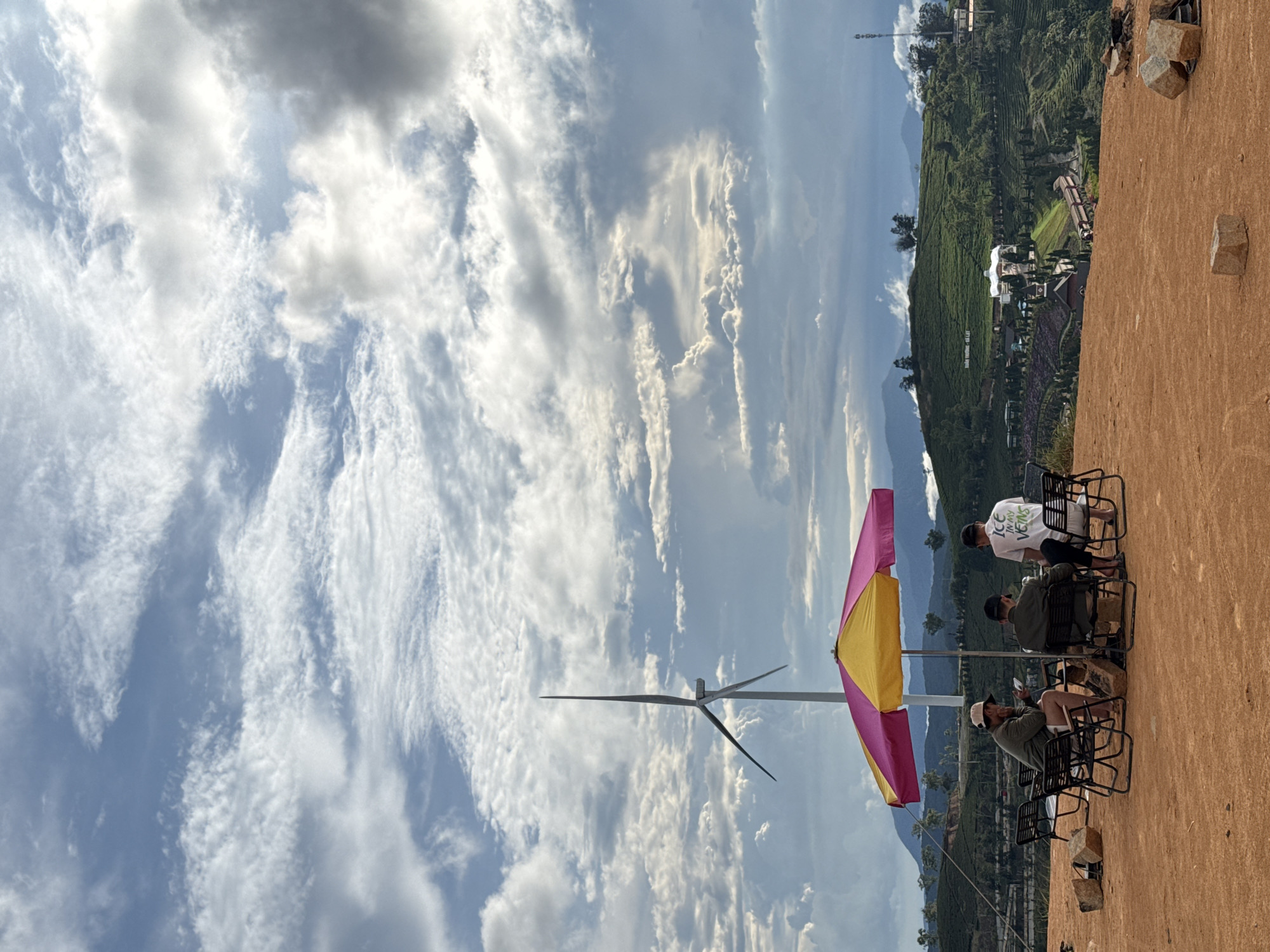 The image shows an outdoor café in what is reported to be Phát Chi, Vietnam. Three people are sitting in black metal chairs at a table shaded by a yellow and pink umbrella. A wind turbine is visible in the distance. The background includes tea plantations on rolling hills, and a sky filled with wispy clouds. The ground is a red-brown color.