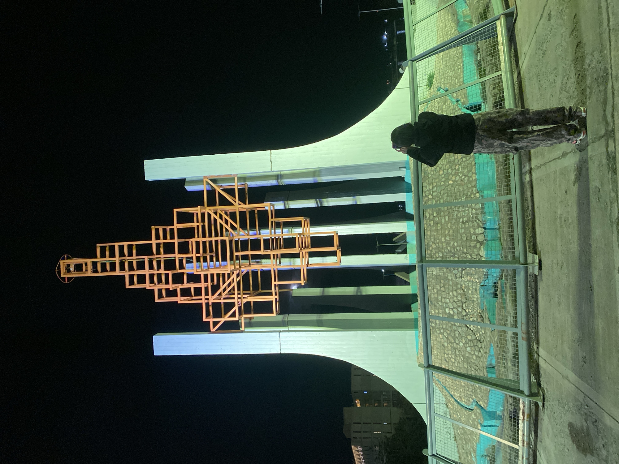 A person is standing on a sidewalk, next to a metal fence and a mural. The person is holding a cell phone, and appears to be taking a photo of a sculpture in the background. The sculpture is made of illuminated wooden frames, and sits atop a large architectural structure. The scene is lit by artificial lights, and a building can be seen behind the sculpture. This is the Monument to the Fallen in Malvinas War in Ushuaia, Argentina.