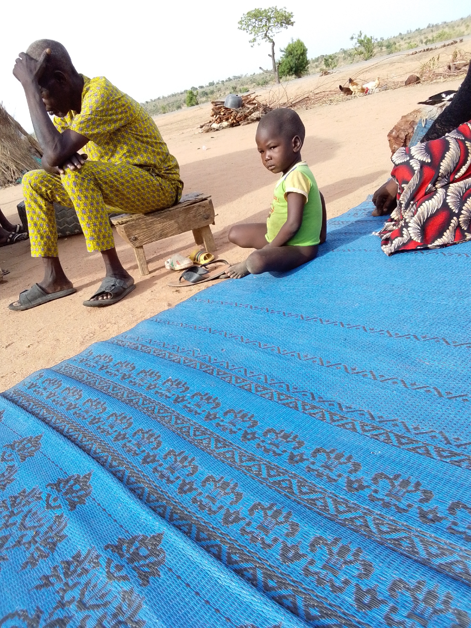 The image depicts an outdoor scene in Mulke Barikin Sayejo, Nigeria, with several people present. A man in a yellow patterned outfit sits on a small wooden stool, his head in his hand. A young child in a green shirt sits on a blue patterned mat. A person wearing a red and white patterned garment sits nearby. A pile of wood and several chickens are visible in the background. The ground is sandy and dry.
