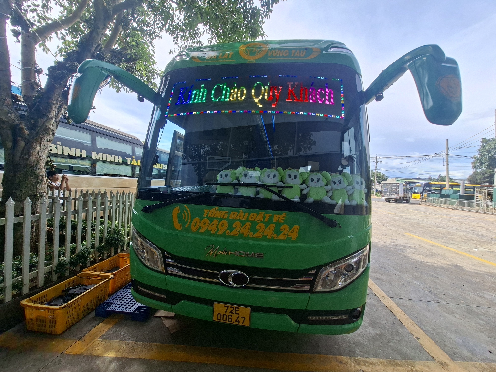 A green tour bus is parked on an asphalt lot, likely in the city of Tân Phú 2, Vietnam, given the context. The bus has a prominent LED display on the front windshield showing the message "Kính Chào Quý Khách" in colorful letters. Below this, a phone number, "0949.24.24.24," and the text "TỔNG ĐÀI ĐẶT VÉ" (booking hotline) are displayed in yellow. A logo that says "MobiHOME" is also visible.
Inside the bus, several green stuffed toys, possibly cartoon characters, are lined up on the dashboard. Two yellow plastic crates containing what appear to be flip-flops are placed to the left of the bus, next to a white metal fence. A person, partially obscured by a tree, is visible behind the fence, seemingly interacting with something they are holding.
The bus has large side mirrors and its headlights are visible. A yellow license plate with the characters "72E 006.47" is affixed to the lower front bumper. In the background, other buses and vehicles are present, along with power lines and trees. The overall scene suggests a transportation hub or a parking area for tour buses.