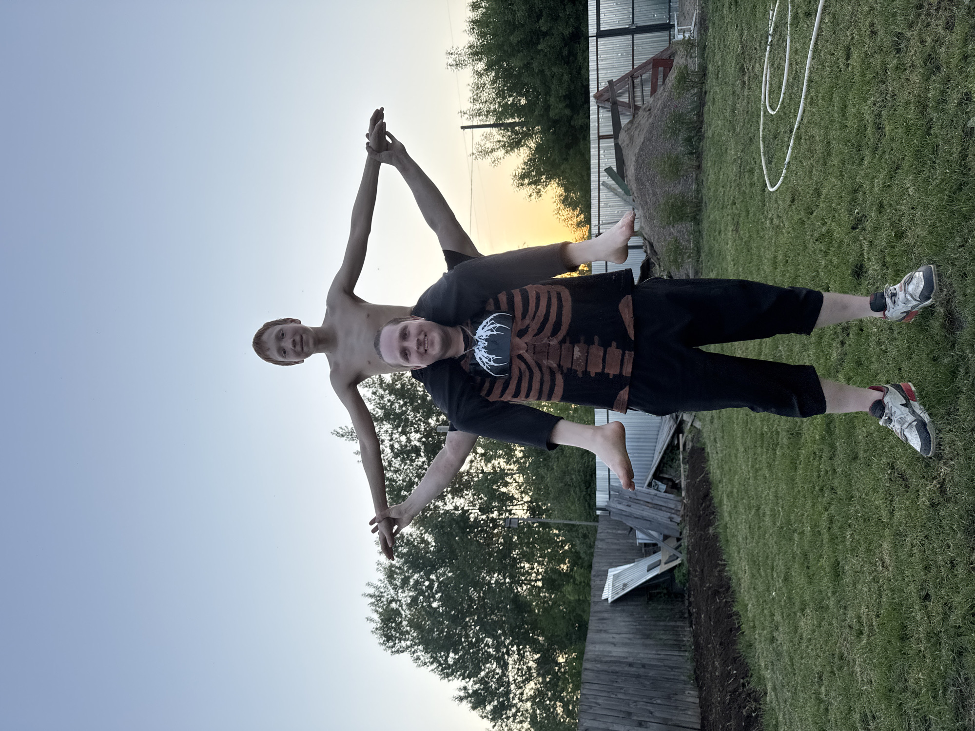 Two males are depicted in an outdoor environment near Chaikovsky, Russia. One male, shirtless, holds his arms upwards to the sky, hands touching. The second male, wearing a black shirt with a skeleton rib cage graphic and black pants, is underneath the first male, supporting him. The background includes trees, grass, a metal structure, and the sky. One of the second male's feet is bare, and the other is in a shoe.