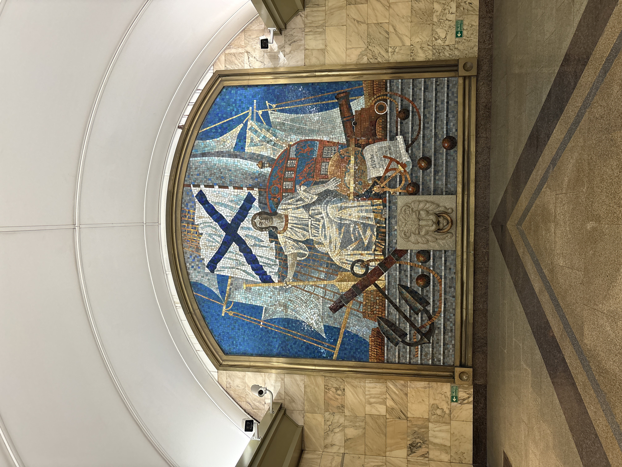 The image shows a large, ornate mosaic depicting a historical naval scene. At the center is a seated figure in white robes, possibly personifying a naval deity or historical figure, holding a globe or astrolabe. Behind the figure are sails and the stern of a sailing ship, rendered in shades of blue and white. To the left and right of the central figure are anchors, cannons, and cannonballs, all rendered in brown and gray tones. A lion's head carving is visible at the bottom center of the mosaic. The mosaic is framed by a golden border and set against a marble-clad wall. Above the mosaic, the ceiling is arched and white, with visible lighting fixtures and security cameras mounted on the wall to the left and right. The floor in front of the mosaic is made of polished stone tiles with a pattern of diagonal lines. A green sign with an arrow pointing left and the word "ВЫХОД" (Exit) is visible on the wall to the left of the mosaic. This mosaic is located in the Narvskaya metro station in Saint Petersburg, Russia, and is dedicated to the history of the Russian Navy.