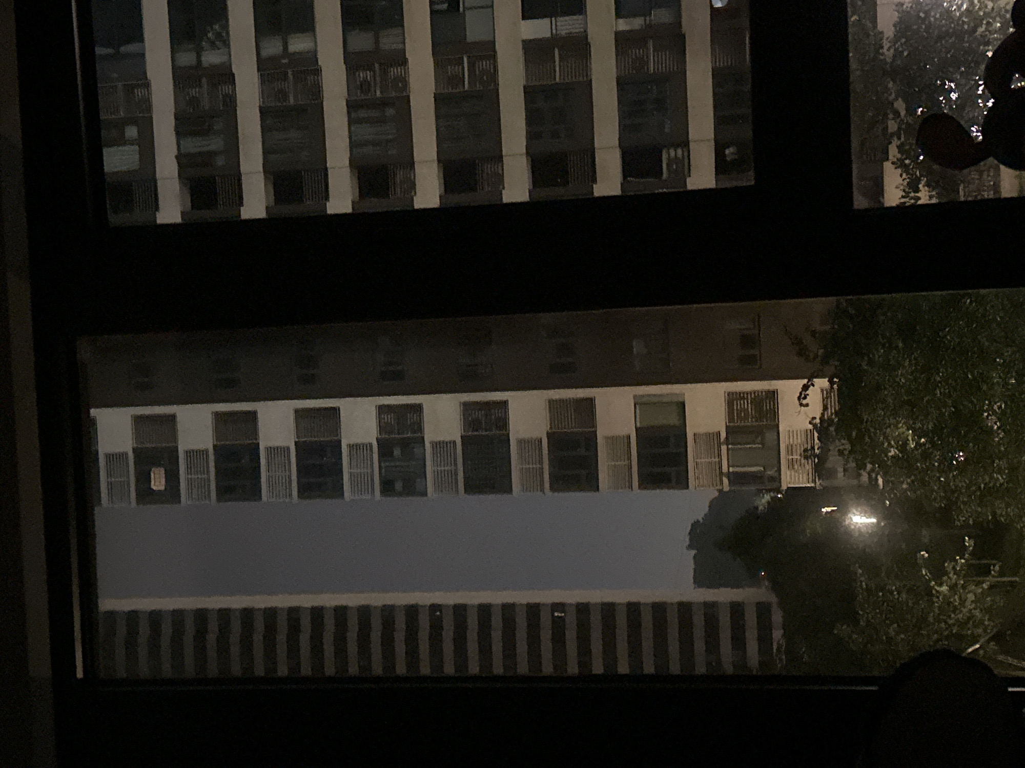 The image captures a nighttime view of urban architecture, likely from a window, looking out at several tall buildings. The scene is dominated by dark silhouettes and artificial lighting.
On the left, a tall, narrow building with a facade of horizontal dark and light bands is visible. To its right, a wider building features a grid of windows. One window on an upper floor of this building has a rectangular, light-colored object within it, possibly a sign or display. Further to the right, another building with a similar grid of windows is prominent. The windows in this building appear darker, suggesting less internal illumination.
The foreground of the image shows dense foliage, likely trees, with some scattered lights reflecting off their leaves, indicating the presence of streetlights or other external light sources. The overall atmosphere is dark and somewhat hazy, typical of a city at night.
The location is identified as the Chongqing High-tech Zone, China.
