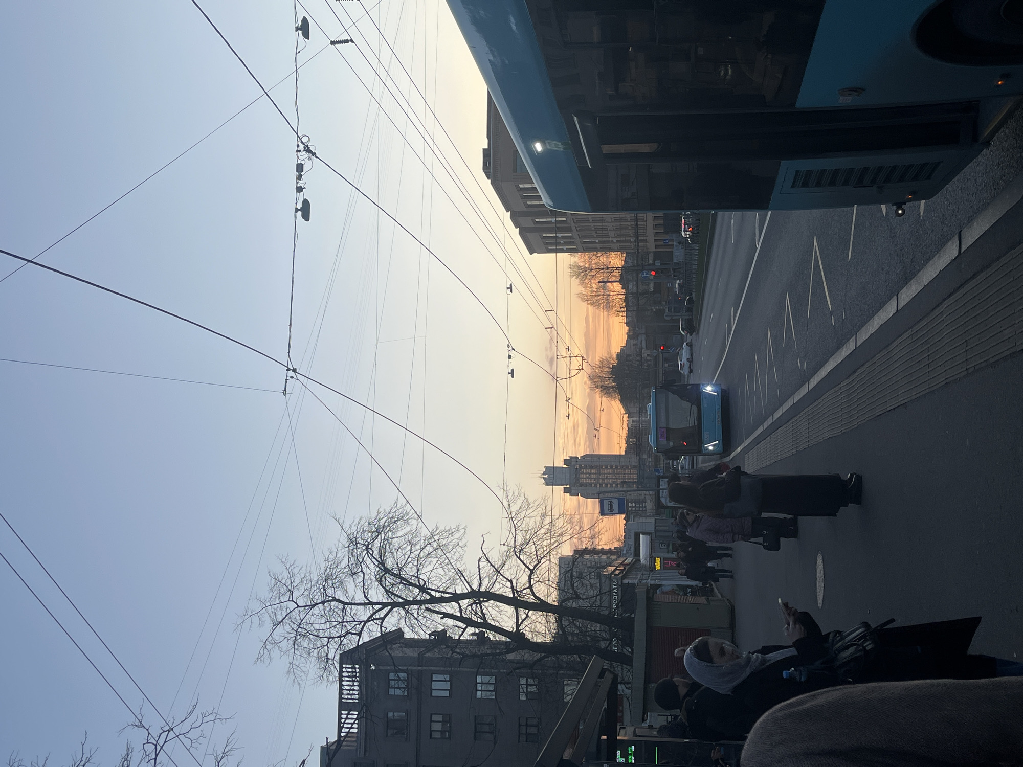 The image is an eye-level, slightly tilted, outdoor urban shot, likely taken from a moving vehicle in Saint Petersburg, Russia. The perspective is towards a street lined with buildings, under a sky transitioning from a pale blue to a warm orange-yellow horizon, suggesting either sunrise or sunset.
To the right of the frame, part of a blue bus is visible, indicating the viewpoint is likely from inside another public transportation vehicle. The street is paved and appears to have a bus lane. A group of people are standing on the sidewalk waiting to board. A woman in the foreground is seated, using a smartphone, and another individual is visible in a hijab.
Overhead are numerous power lines crisscrossing the sky. Buildings of varying heights are visible, some with a Soviet-era architectural style. The cityscape is moderately dense, with signs of commercial activity. The absence of leaves on the trees suggests a colder season. The scene is composed of multiple elements indicating a typical urban environment in Saint Petersburg.