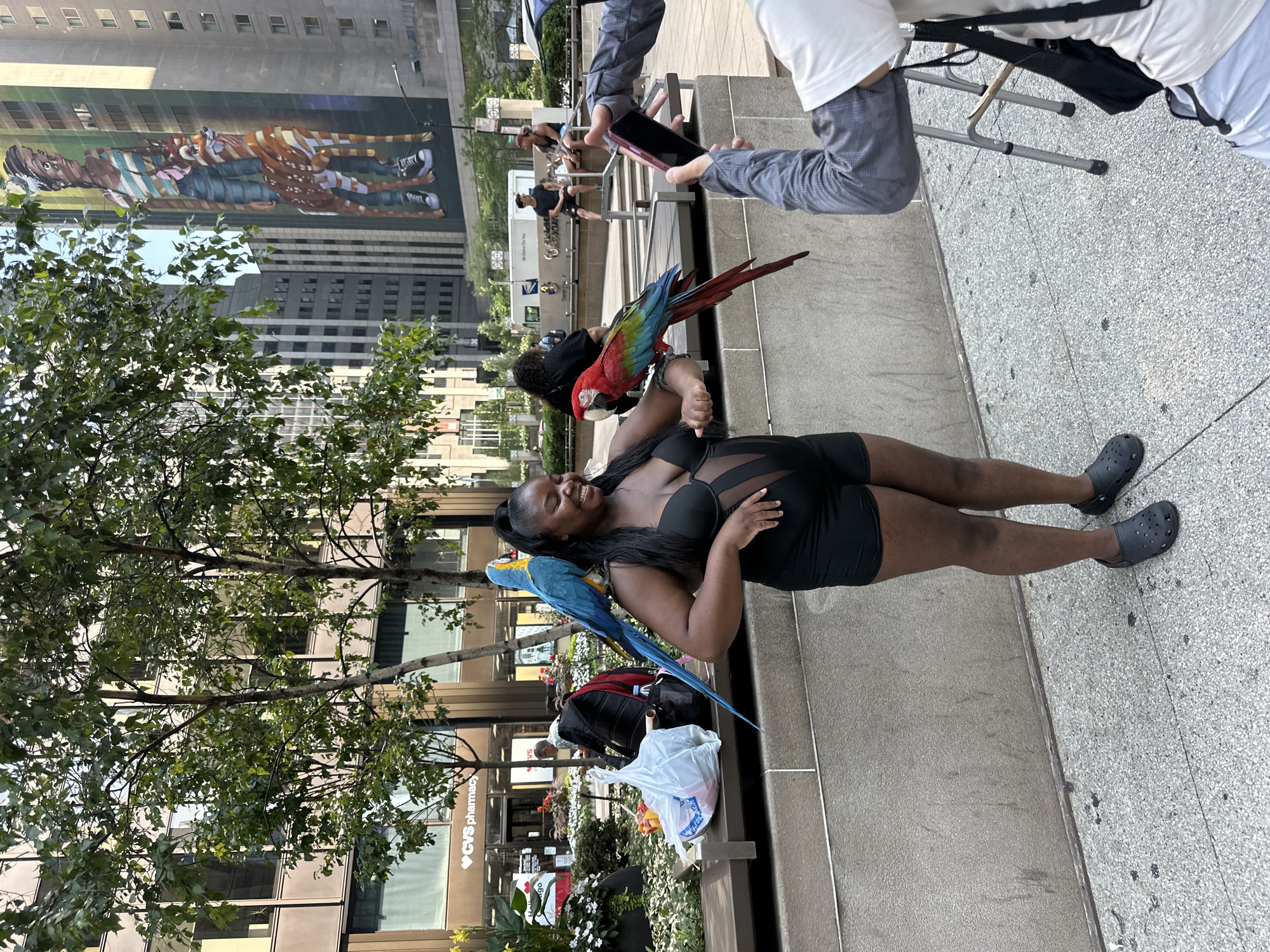 In Chicago, Illinois, United States of America, a Black woman stands outdoors on a paved walkway. She is wearing a black bikini top and black shorts, with black crocs on her feet. Two parrots are perched on her. A blue and yellow macaw is on her left shoulder, and a red, green, and blue macaw is on her right arm. The woman is smiling and looking to her right. In the foreground, a person's hands are visible holding up a mobile phone, as if taking a picture or video. Behind the woman, there is a stone wall and a bench. On the bench, there is a plastic bag filled with items. Further in the background, there are trees, buildings, and other people. One building has a large mural on its side depicting a young woman and a deer-like creature. A sign for CVS pharmacy is visible on another building. A white delivery truck is also visible in the background.