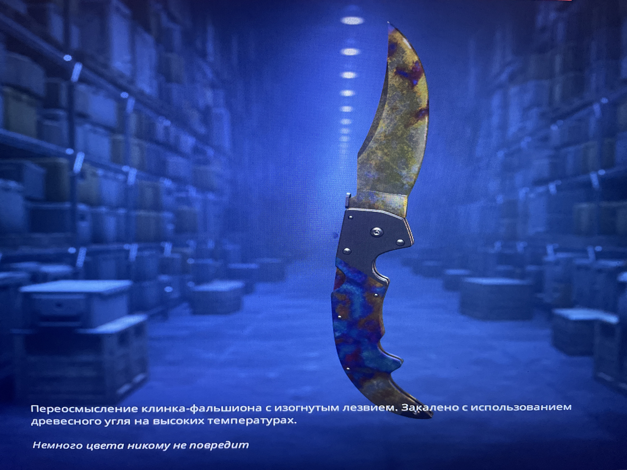 The image shows a digitally rendered Karambit knife with a unique "Fade" skin, characterized by its vibrant, iridescent blue, purple, and yellow hues, set against a dark, blue-toned background resembling a warehouse or storage facility. The knife is the central focus, with its curved blade and intricately patterned handle prominently displayed. Text in Russian is present at the bottom of the image, describing the knife's properties: "Reimagining of a falchion with a curved blade. Hardened using charcoal at high temperatures." and "A little color will not hurt anyone." The background features rows of stacked boxes or containers, dimly lit and out of focus, suggesting an industrial or logistical environment. There is no indication of the city of Kołobrzeg, Poland, or any specific real-world location.