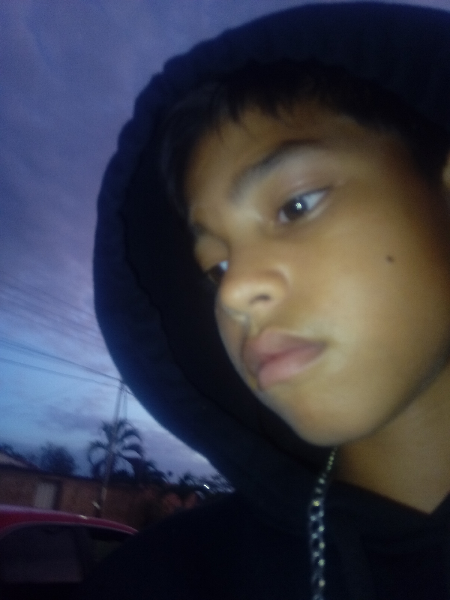 A close-up photograph shows a young person with brown skin and short, dark hair wearing a black hooded sweatshirt. The hood is up. A silver chain is visible around the person's neck. The background is blurry, but a red car and a palm tree can be discerned, and the sky is a mix of purple and blue, indicating either dusk or dawn. The location is Juan Griego, Venezuela.