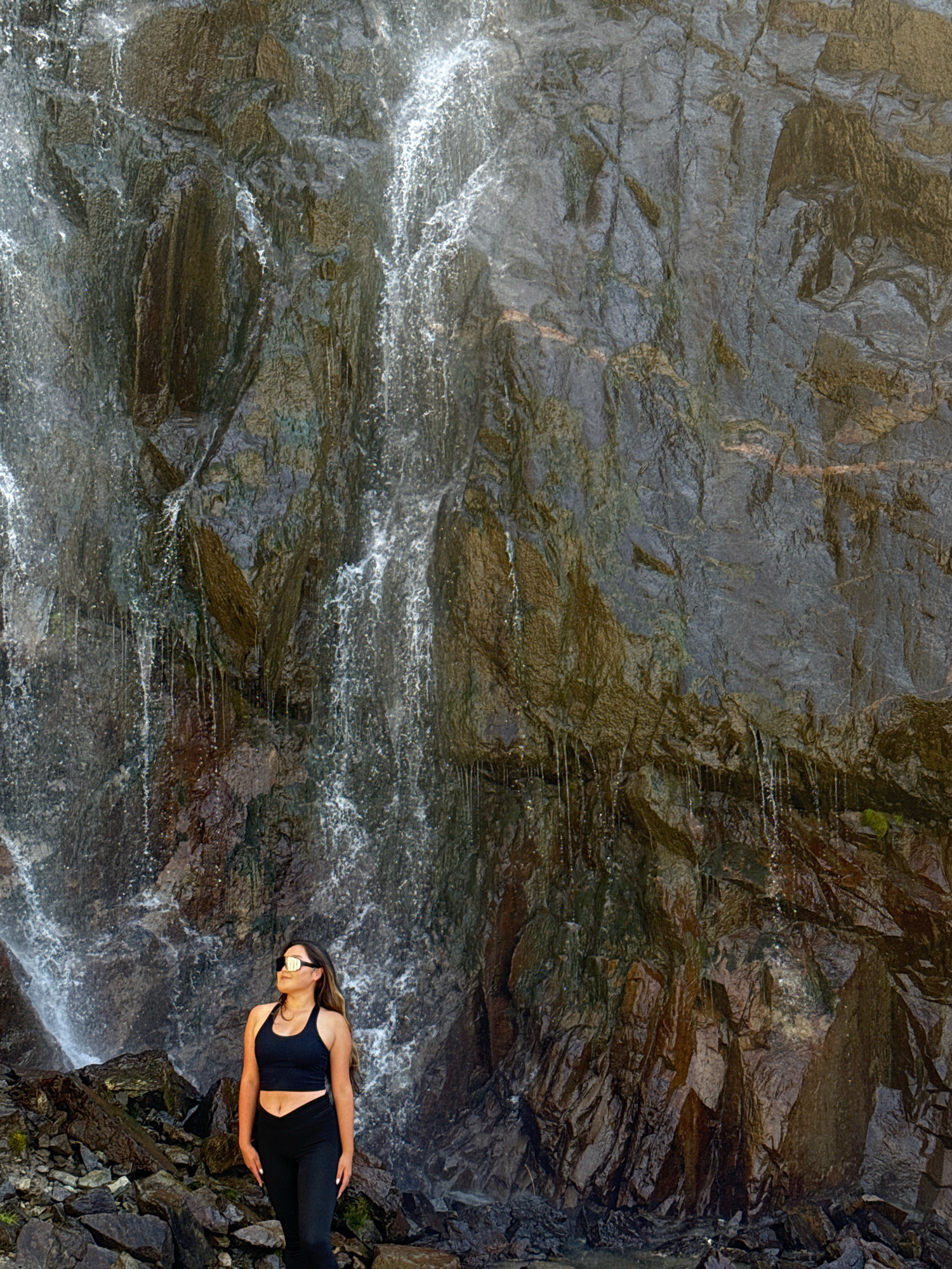 A full-body, eye-level shot shows a woman standing in front of a waterfall. The woman is facing right with her head tilted up, looking towards the top of the waterfall. She has long, dark hair and is wearing a black sports bra, black leggings, and sunglasses. The waterfall cascades down a rocky cliff face, with water spraying and dripping. The rocks are dark and wet, with some moss and vegetation visible. The foreground is filled with dark, jagged rocks. The image appears to be taken outdoors in a natural environment. The location is given as Unknown, Kyrgyzstan.