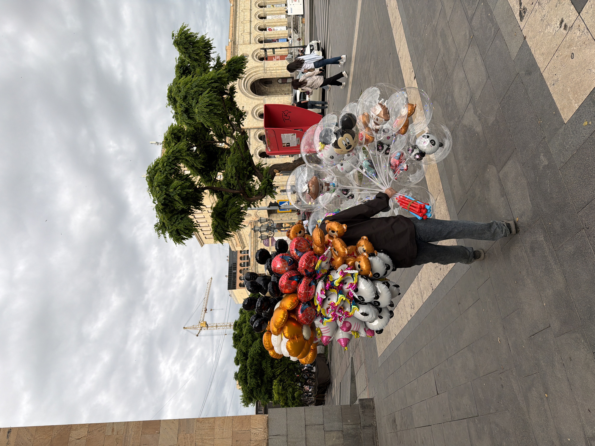 A street vendor in Yerevan, Armenia, stands on a paved public square holding a large assortment of helium balloons. The balloons are a mix of clear inflatables with toys inside and character-themed designs such as Mickey Mouse, pandas, Spider-Man, and bears. The vendor wears a dark jacket and denim jeans. Behind the vendor are pedestrians walking near a red box, and a stone building with arches. A tree is to the left of the red box. In the background, a construction crane is visible against a cloudy sky.