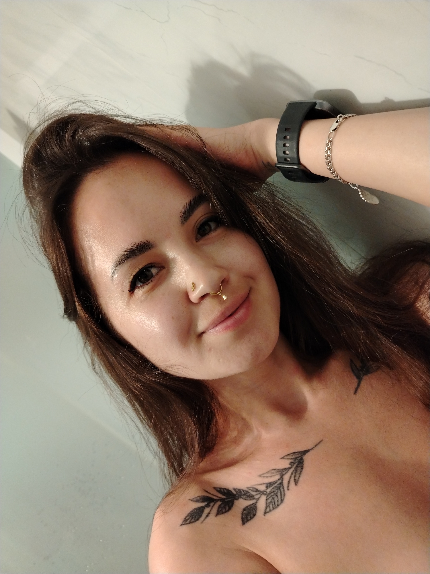 A selfie of a fair-skinned young woman with long, brown hair, a gold nose ring, and leaf tattoos on her chest and shoulder. She is wearing a black smartwatch and a silver bracelet on her left wrist. She is indoors. The location is unspecified; Noviy, Russia, is not discernible.