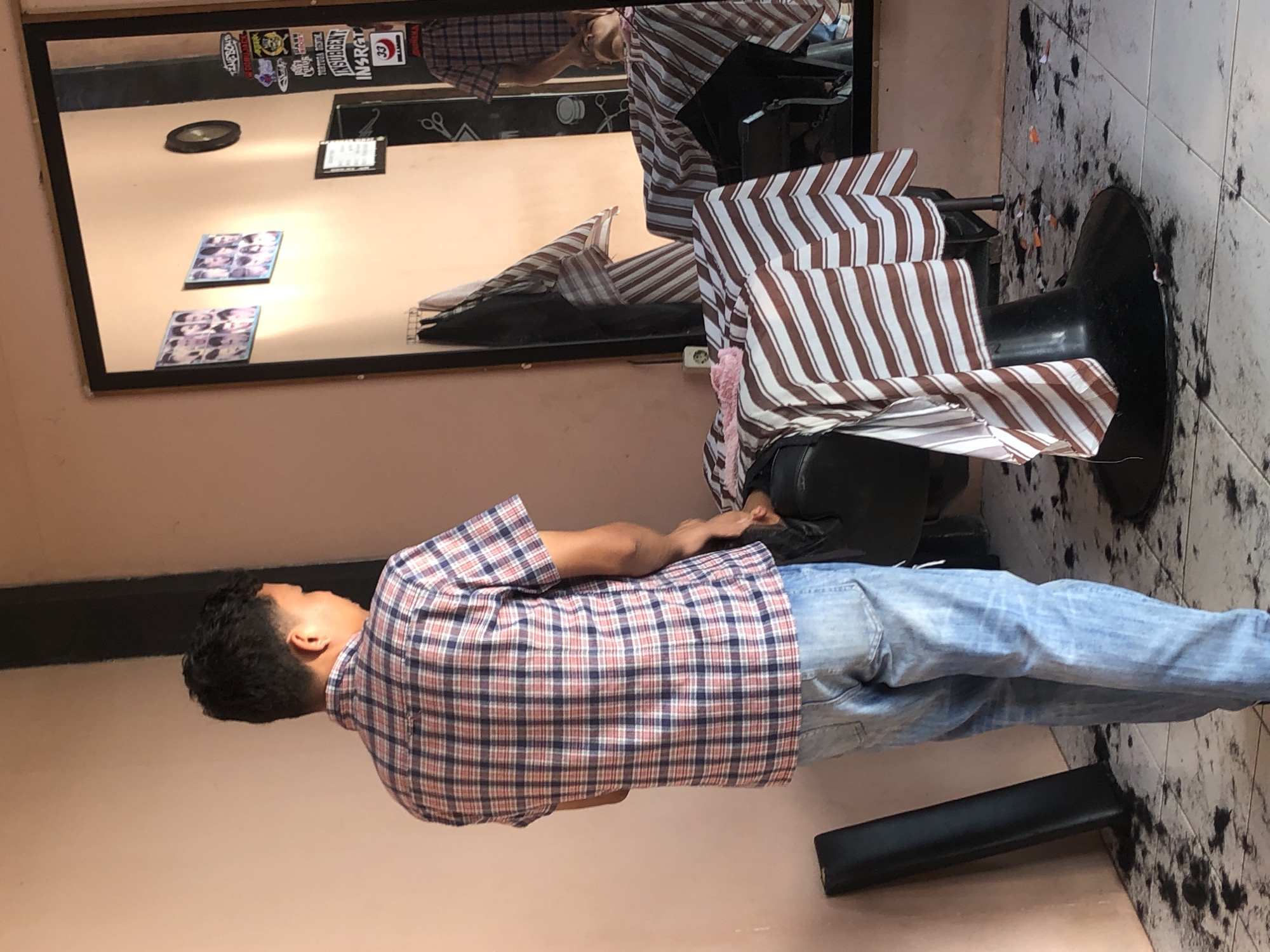 A person wearing a plaid shirt and blue jeans sits in a barber chair covered with a striped cape. The barber shop is in Pulorayeuk, Indonesia. Hair clippings are scattered on the tiled floor. A mirror hangs on the wall, reflecting the person in the barber chair, as well as some items on the wall. These include stickers, pictures and a clock. A black footrest is visible near the chair.