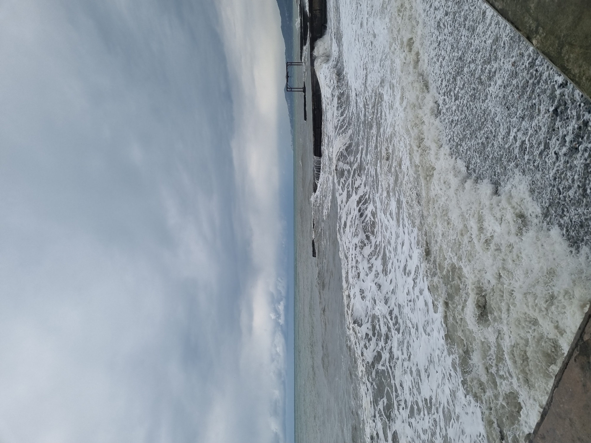 The image depicts a seascape, potentially taken in the city of Sochi, Russia. A turbulent sea is visible on the right side of the image, with waves crashing against a concrete structure. A pier extends into the water. The sky is overcast with varied cloud formations. The horizon line is situated midway in the image, separating the sky and sea. A coastline with possibly mountainous terrain is seen in the distance.