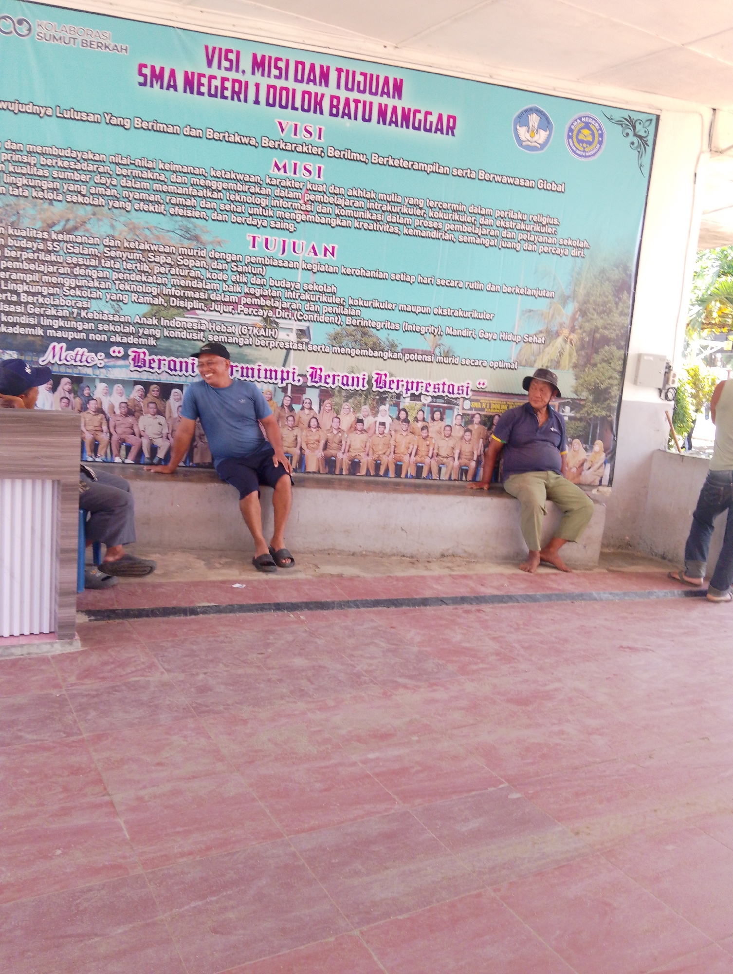 The image shows a group of people gathered in front of a large banner at SMA Negeri 1 Dolok Batu Nanggar, located in Serbelawan, Indonesia. The banner displays the school's vision, mission, and objectives. In the foreground, several men are sitting on a concrete bench. One man in a blue t-shirt and a black cap is smiling and leaning on the bench. Another man in a dark blue polo shirt and a brown hat is also seated, looking to his left. Two other individuals are partially visible, one sitting on a blue chair and another standing. The background features a large banner with text and images, likely representing the school's community and educational activities. The overall environment appears to be an outdoor gathering space, with a tiled floor and a building structure to the right.