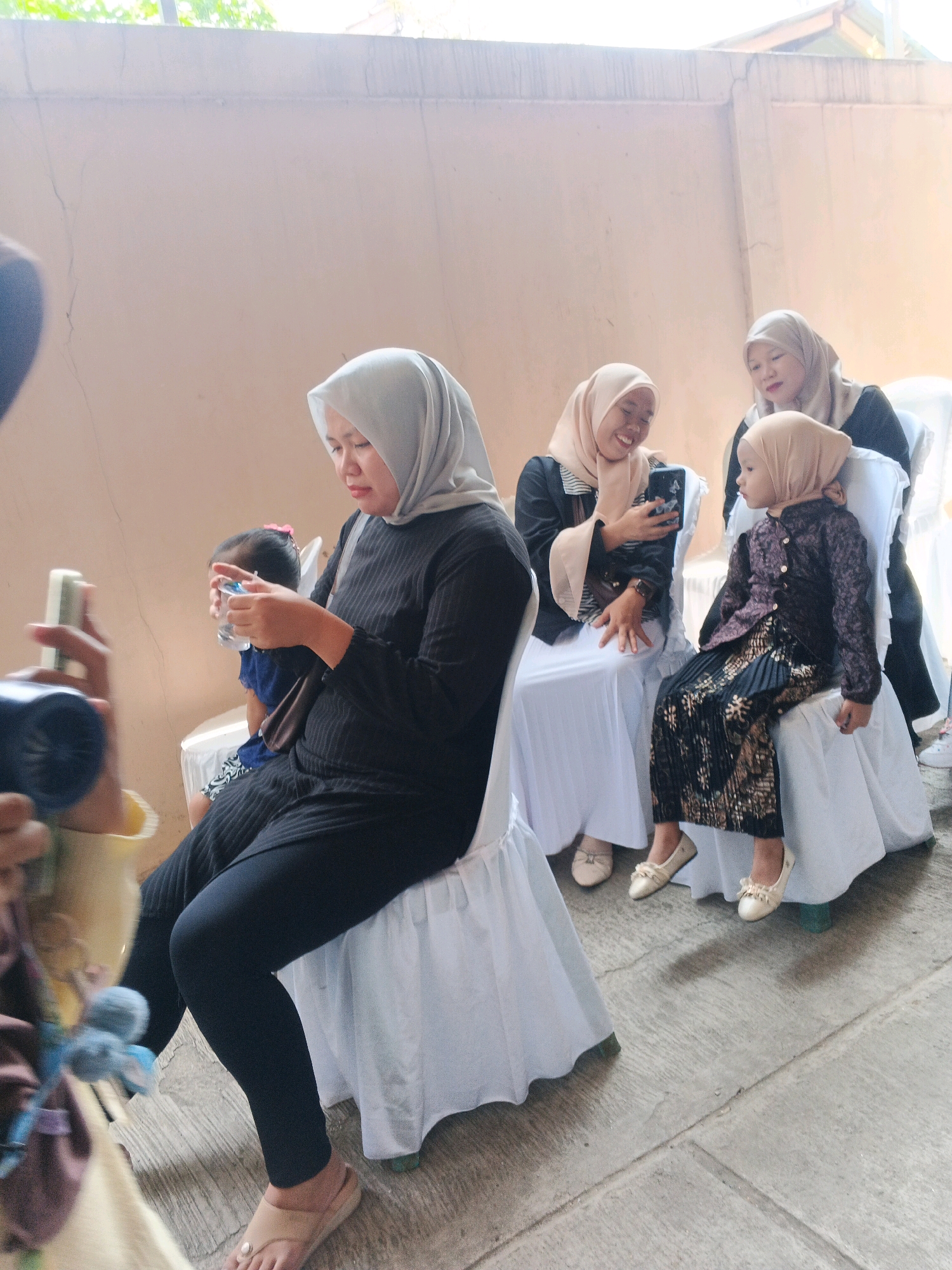 In Kadu, Indonesia, an outdoor gathering is taking place near a light-colored wall. Four individuals, all wearing hijabs and modest attire, are seated on white-covered chairs. A woman in a black dress and light grey hijab is in the foreground, looking down at a small bottle of water in her hands, with a child in a blue shirt partially visible behind her. To her right, another woman in a peach hijab is smiling and looking at a mobile phone, holding it up as if taking a photo. Next to her, a young child in a patterned black and gold outfit and a peach hijab is seated, facing to the left. Behind them, a fourth woman, also in a peach hijab, is seated, her body partially obscured. The ground is paved with concrete slabs. The lighting suggests it is daytime, possibly afternoon.