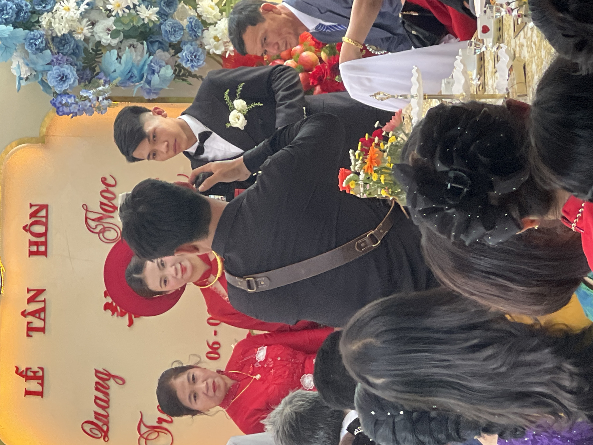Captured indoors in Bình An, Vietnam, a wedding ceremony is in progress. A man in a black suit and bow tie is visible, along with another individual in a suit who may be an elder. A photographer with a shoulder strap focuses on the bride and what appears to be her mother, both dressed in red. White and blue floral arrangements adorn the backdrop, which bears red script and appears to be a celebratory banner. The scene includes a table with a white cloth and various decorative items. Several spectators stand in the foreground.