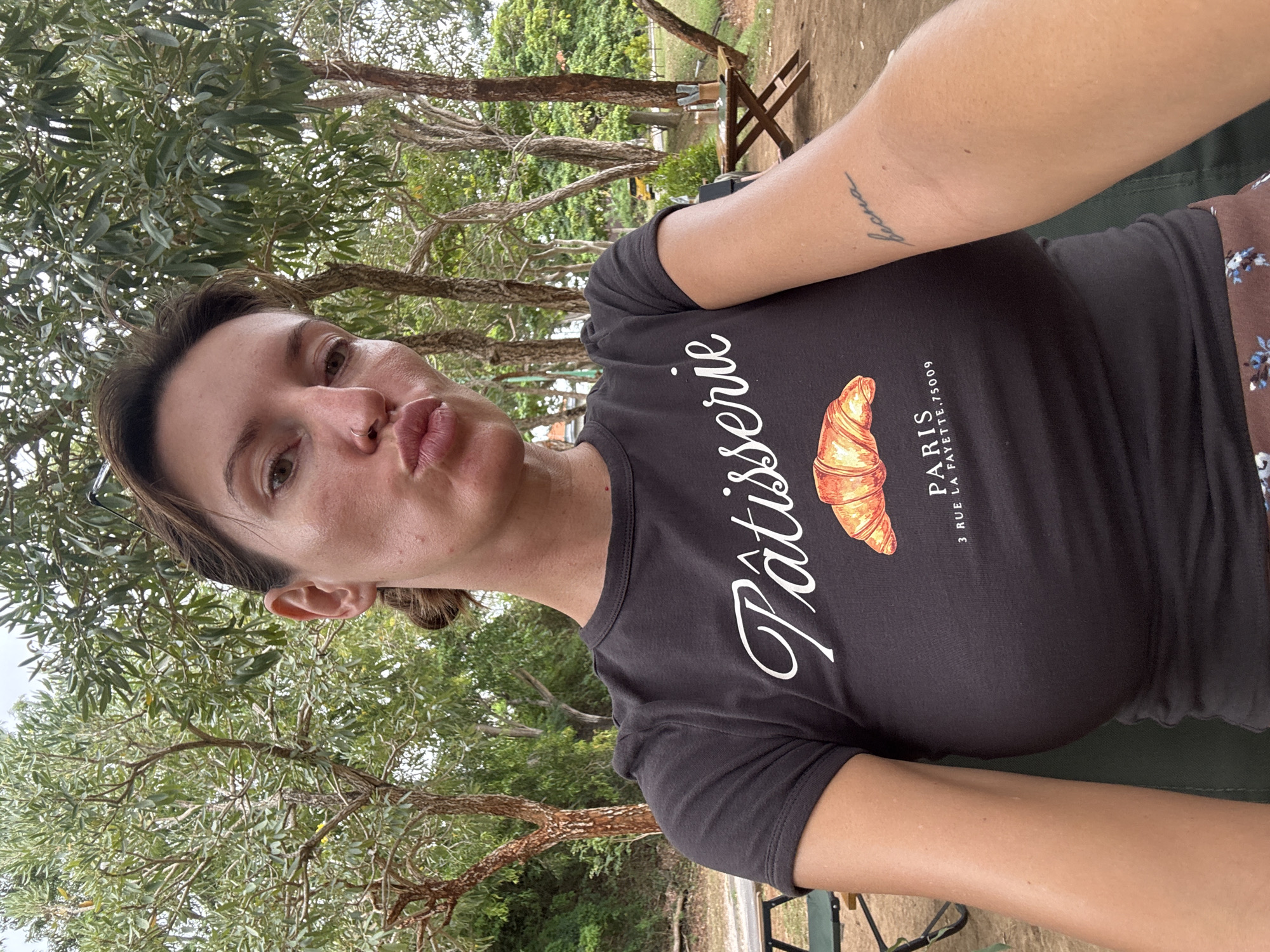 A woman is taking a selfie outdoors. She is wearing a black t-shirt with a graphic of a croissant and "Pâtisserie" written in white script. Her hair is pulled back, and she is puckering her lips. A small tattoo is visible on her left forearm. The background shows trees and foliage, suggesting a park or natural setting. In the distance, a table and chairs can be seen, along with a yellow vehicle. The overall environment is lush and green, indicative of a tropical climate. While the image is tagged as being in Kuta Selatan, Indonesia, there are no specific landmarks or details within the image that definitively confirm this location.