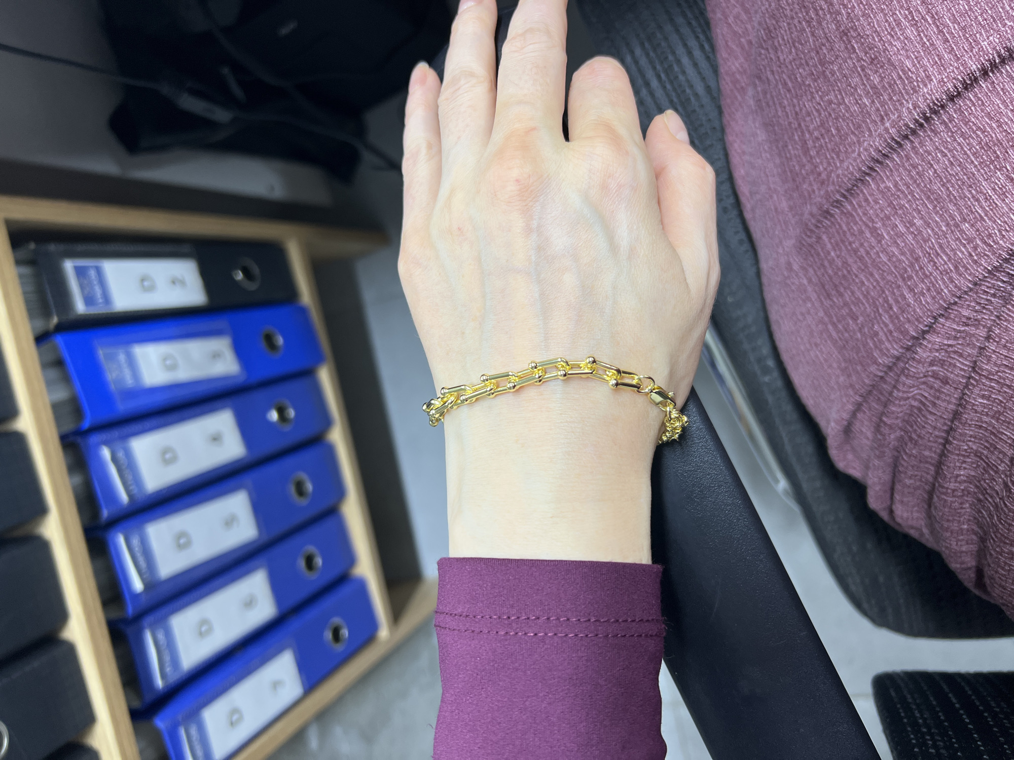 A close-up, eye-level, medium shot shows a person's left arm and hand, adorned with a gold bracelet, resting on a dark chair. The person is wearing a dark purple long-sleeved shirt. In the background, a wooden shelf unit holds several blue and black binders. The scene appears to be indoors, likely in an office setting in Shahrixon, Uzbekistan. The lighting is dim, with a light source illuminating the arm and hand.