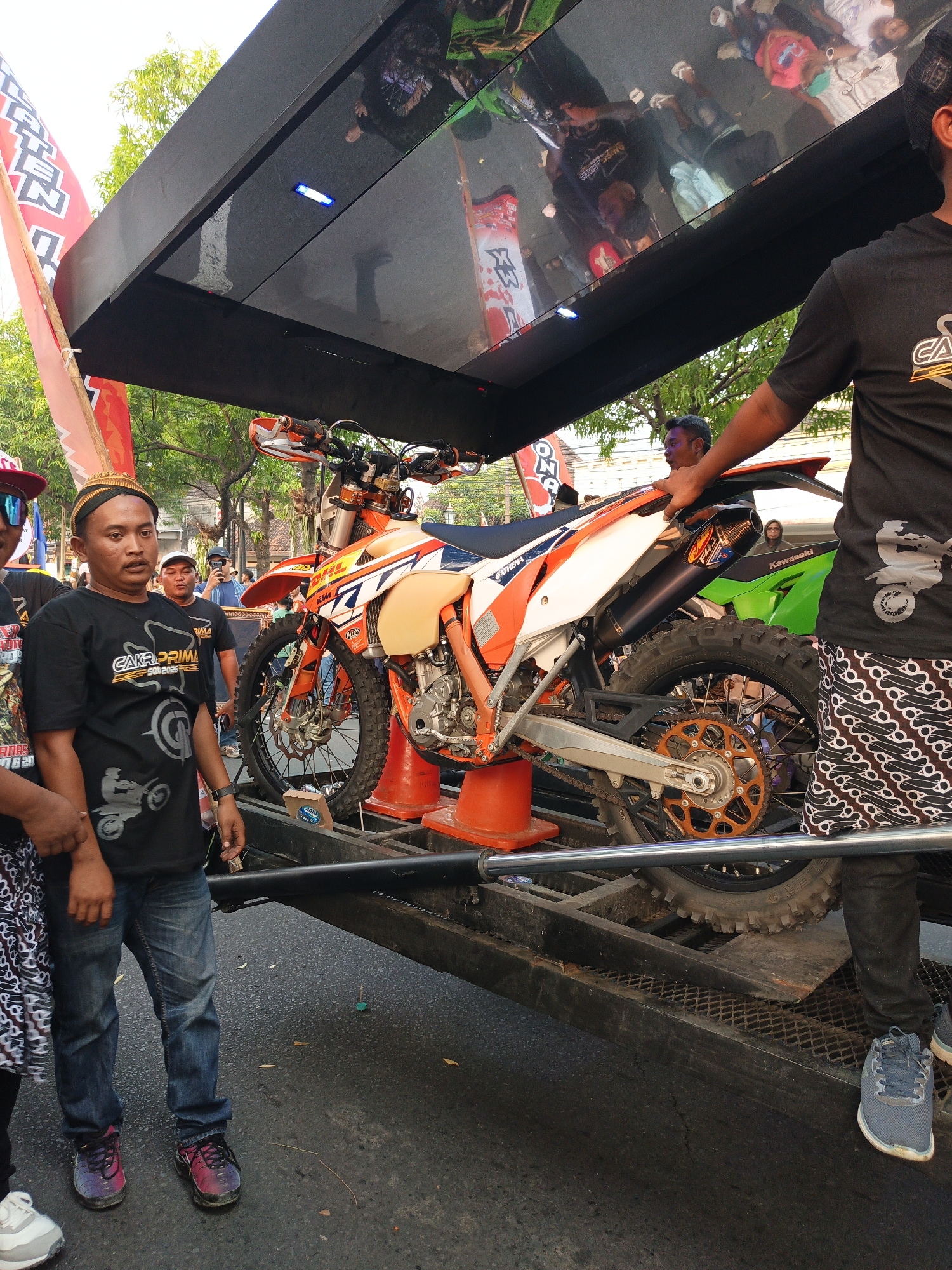 In Gayamprit, Indonesia, a black, elevated structure with a reflective surface is positioned above an orange and white dirt bike loaded onto a trailer. The structure's surface reflects a crowd of people, some holding flags. A blue LED light is visible on the underside of the structure. The dirt bike, a KTM, is a prominent subject, positioned on the trailer with its front wheel slightly elevated by orange cones. People are gathered around the trailer, some looking at the bike, others engaged in conversation. A man in a black t-shirt and jeans stands near the front of the bike, while another man in a similar t-shirt is visible to his left. The environment suggests an outdoor event, with trees and a paved surface visible.