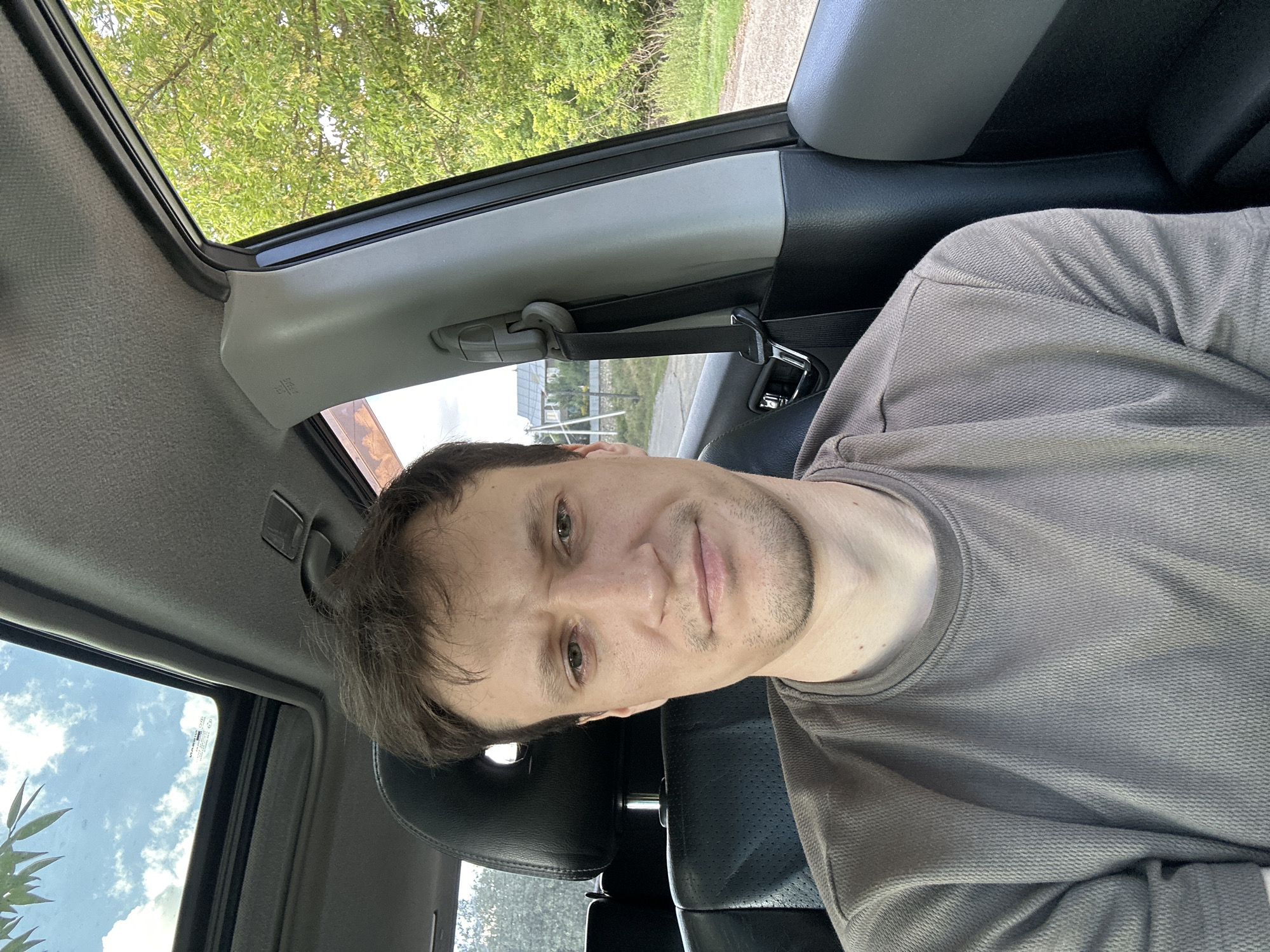 The image shows a man sitting in the driver's seat of a car, facing the camera. He has short brown hair and is wearing a grey t-shirt. The car has a sunroof, which is open, allowing a view of the sky and some trees. The background outside the car is blurry but appears to be a rural or suburban setting with trees and a house visible. The man's expression is neutral. The interior of the car is visible, including the leather seats and the dashboard. There is no specific information about the location within Makiv, Ukraine, other than it is implied by the prompt.