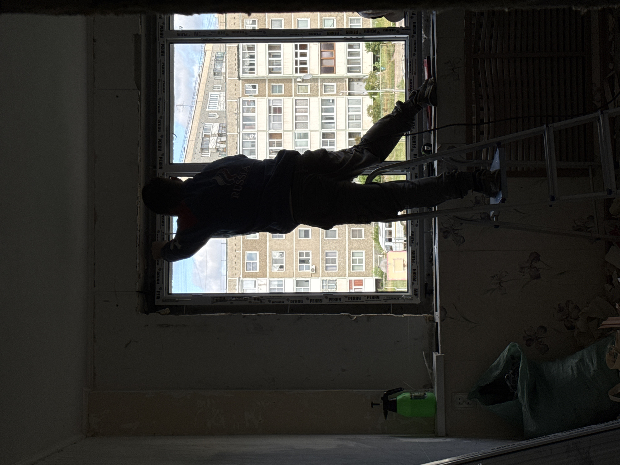 A person, silhouetted against a bright window, stands on a ladder inside a room undergoing renovation. The individual, wearing a dark hooded jacket with "RUSSIA" printed on the back, dark pants, and work boots, is actively engaged in work near the window frame. Tools and materials, including a green spray bottle and a large green bag, are visible on the floor and a windowsill. The window looks out onto a multi-story residential building characteristic of Soviet-era architecture, suggesting an urban environment. The presence of peeling wallpaper, debris on the floor, and exposed construction materials indicates a renovation or construction site. The overall scene depicts an interior workspace in a Russian city, likely Chaikovsky, given the context provided.