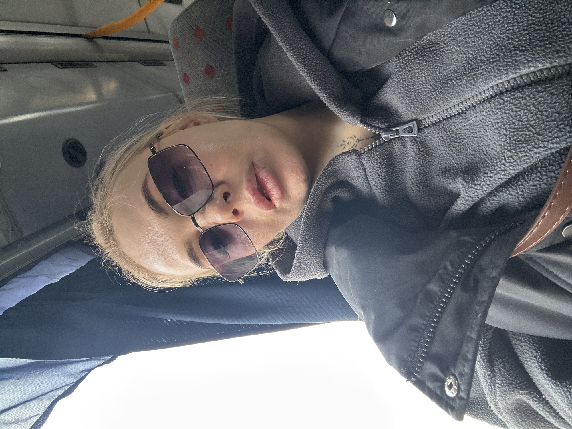 A young woman with blonde hair and fair skin is wearing dark sunglasses and a gray jacket. She is looking at the camera, possibly inside a bus, as indicated by the seating and interior details in the background. The location is inferred as Megion, Russia, based on the image context, but no specific landmark is visible to confirm this. A visible tattoo is on her upper chest.
