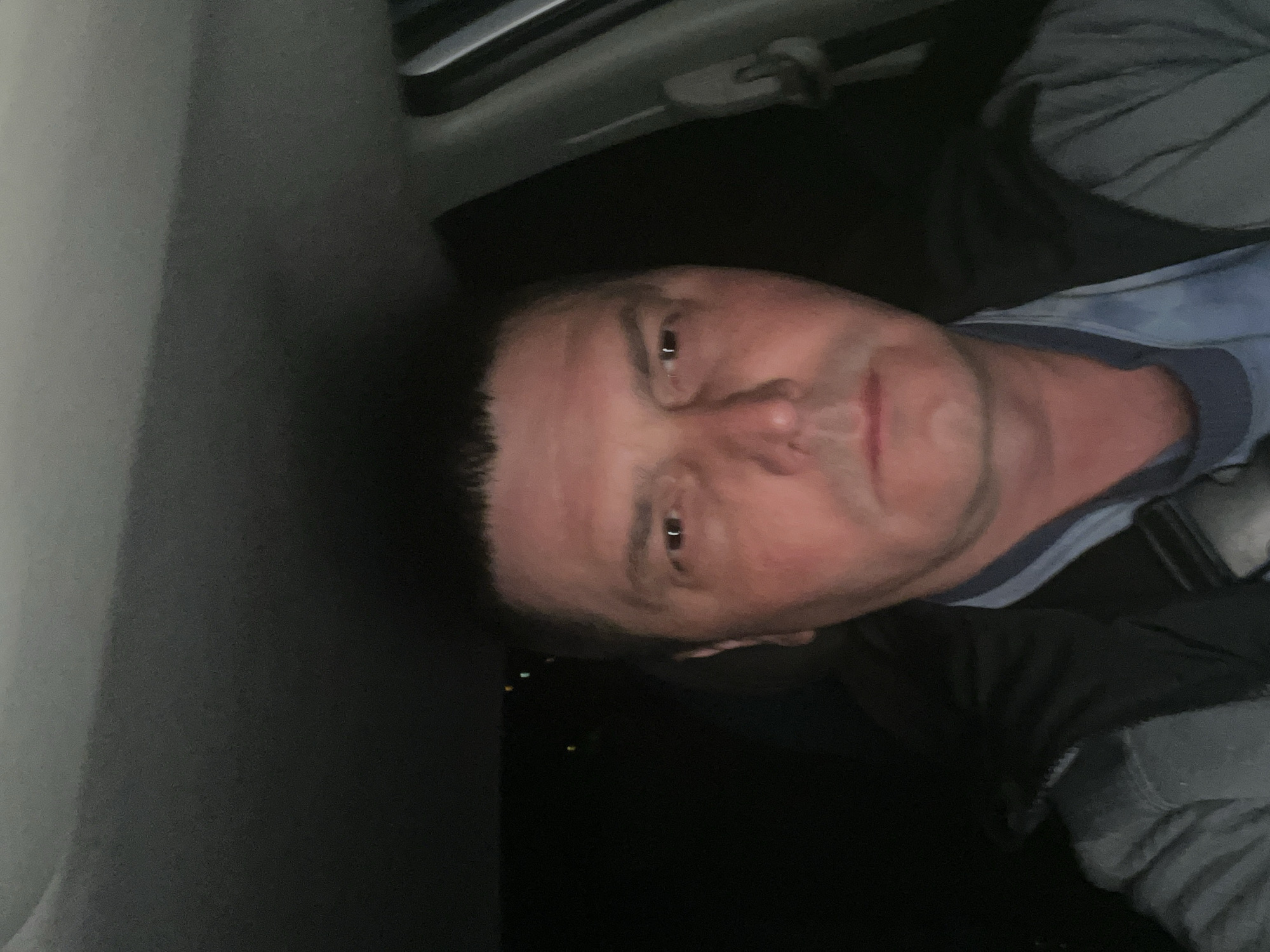 The image is a close-up portrait of a man, appearing to be taken in a car. The man, who seems middle-aged with short dark hair, is positioned with his face and upper torso visible. He is looking directly towards the camera with a neutral, slightly tired expression. His face is well-lit, showing a fair complexion. He is wearing a light blue t-shirt with a subtle pattern.
To the left of the man, a gray interior panel of the vehicle is visible. Above his head, parts of the car's ceiling or headliner, along with a handle or clip, are discernible. The area to his right and behind him is predominantly dark, indicating that the photo was taken at night or in very low light conditions. Small, faint specks of light are visible in the dark background, potentially distant lights or reflections. No discernible text is present in the image.