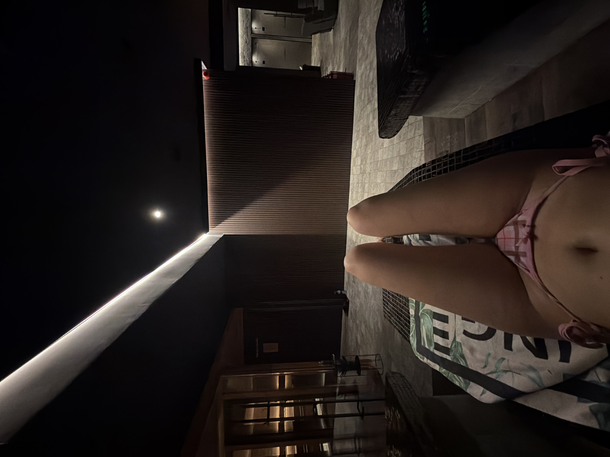 The image shows a person in a pink bikini sitting on a patterned towel in a room with dark walls and a bright light strip along one wall. The room has a wooden wall paneling and tile floors. A shelf-like structure is visible in the lower part of the image. The location is in Sofia, Bulgaria.