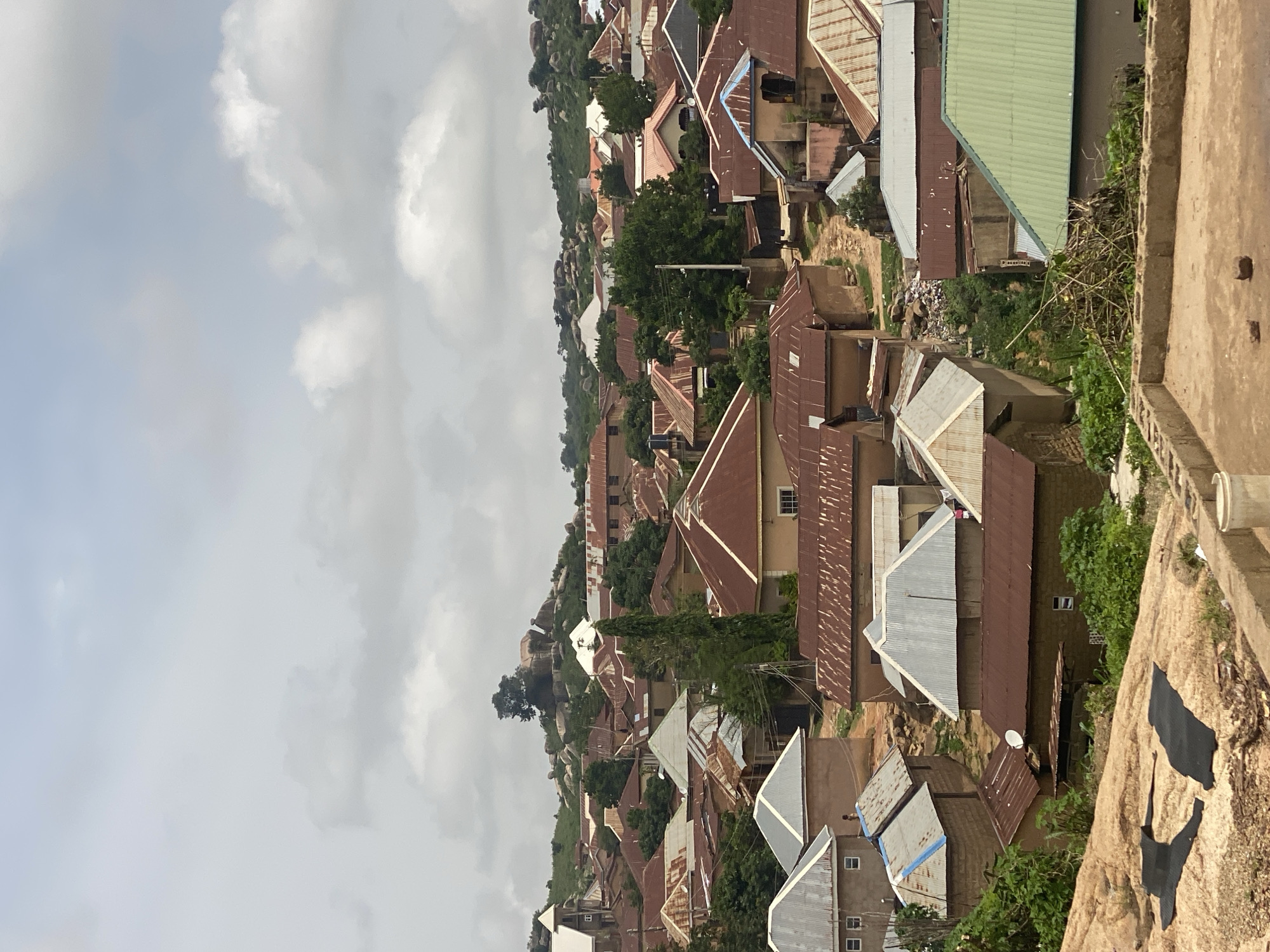 This image captures a sprawling hillside settlement in Jos, Nigeria, characterized by a dense collection of buildings with predominantly corrugated metal roofs in various states of repair. The architecture suggests a mix of residential and possibly small commercial structures, densely packed together along the undulating terrain. Green trees are interspersed throughout the settlement, adding a touch of nature to the urban landscape.
In the foreground, a rough, textured surface, possibly rock or unpaved ground, is visible, with some dark, irregular shapes affixed to it. This area appears to be an elevated viewpoint overlooking the town. The sky is overcast with a significant presence of clouds, suggesting a potentially humid or rainy day, or simply a cloudy sky typical of the region. The time of day is not explicitly clear, but the diffused light indicates it's likely daytime, perhaps mid-morning or afternoon.
The scene depicts a lived-in community, with clothes hanging out to dry visible on some buildings, hinting at the daily life of its inhabitants. The density of the settlement and the rugged landscape suggest a challenging but established living environment. No explicit activities or events are prominently depicted, but the overall impression is one of a functional, perhaps developing, urban area nestled within a natural, rocky environment. There is no visible text in the image.