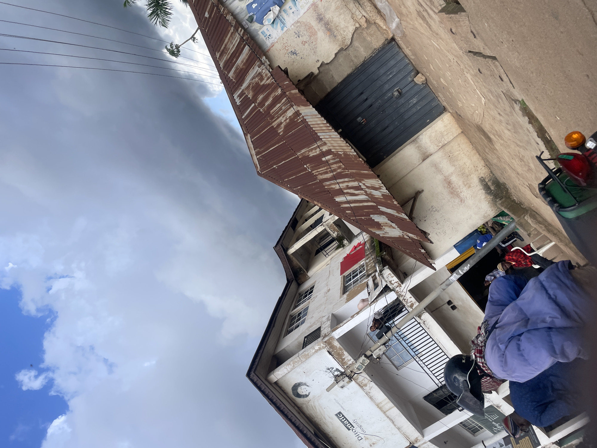 This image captures a street scene in Gura Top, Nigeria. In the foreground, a person wearing a blue puffer jacket and a black helmet with a red and white scarf is seen from the back, sitting on a motorcycle. Behind them, a portion of the motorcycle's rear with red and orange lights is visible. To the left, another person in a dark blue jacket is partially visible.
The middle ground of the image features two adjacent buildings. The building on the left is a multi-story structure with white walls, balconies, and windows. A mural of a face is painted on its side, along with text that appears to read "Ditomatic Otography". Red clothing is hung on a balcony on the second floor. A utility pole with electrical wires stands in front of this building.
The building on the right has a weathered, rusty corrugated metal roof and an off-white, somewhat dilapidated wall. A large, dark, metal shuttered door is visible on this building, with a padlock. Posters or advertisements are affixed to the wall next to the door, one of which displays a blue jacket with a white letter "P".
In the background, a dramatic sky with dark, heavy clouds dominates the upper portion of the image, suggesting impending rain. Electrical wires are strung across the sky. A palm tree is visible to the far right, partially obscured by the roof of the building. The ground in the foreground and middle ground appears to be a dirt or concrete surface. People are visible on the balconies of the building on the left and near the entrance of the building on the right, suggesting a populated urban environment.