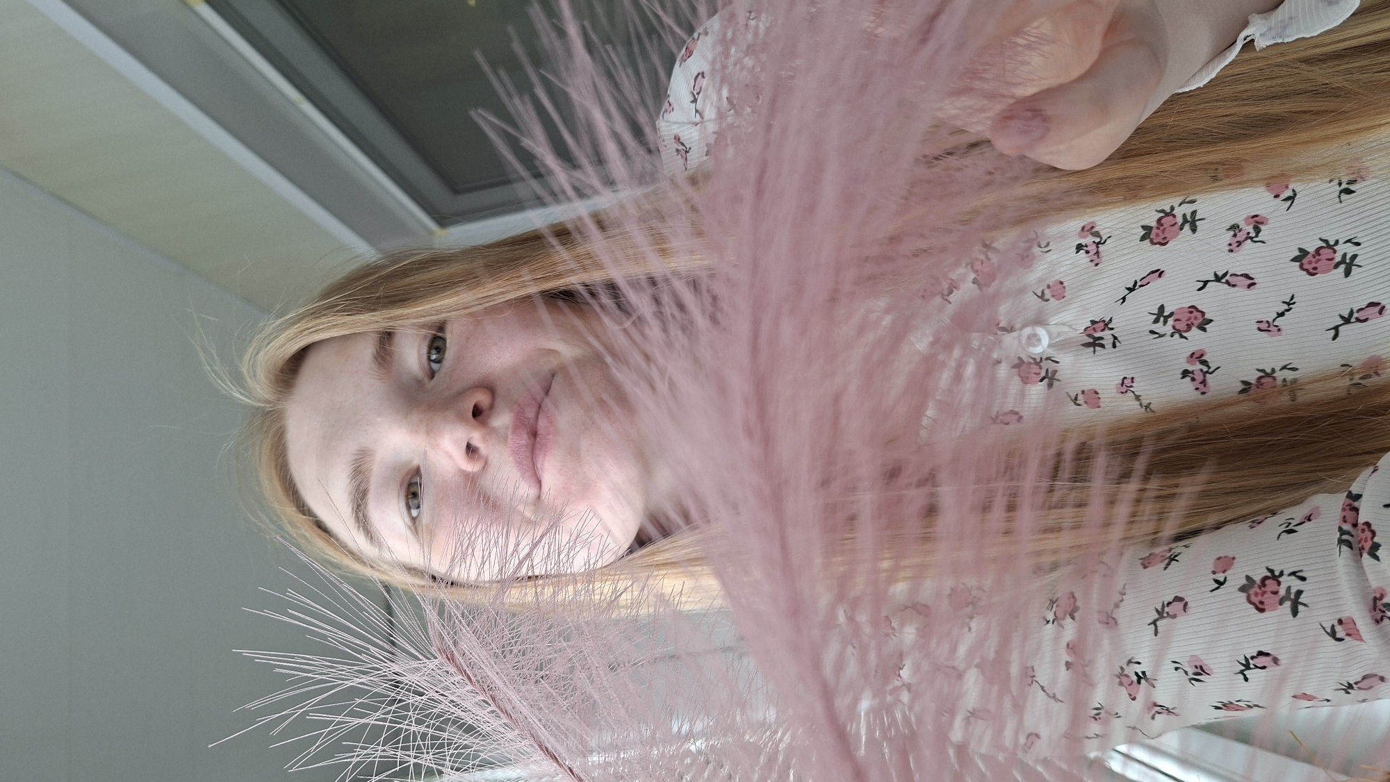 A close-up, high-angle shot captures a young woman from Chaikovsky, Russia, looking up towards the camera. The image is taken at an angle, with her head rotated so her right eye is higher than her left. Her blonde or light brown hair cascades around her, partially covering her white top adorned with a pattern of small pink and red flowers and green leaves. She holds a cluster of delicate, fluffy pink pampas grass in her left hand, which extends across the frame, creating a soft, blurred foreground. Her eyes are light brown, and she has a hint of a smile. The background is simple and out of focus, suggesting an indoor setting with possibly a window or a light-colored wall. The overall mood is soft and gentle. No visible text is present.