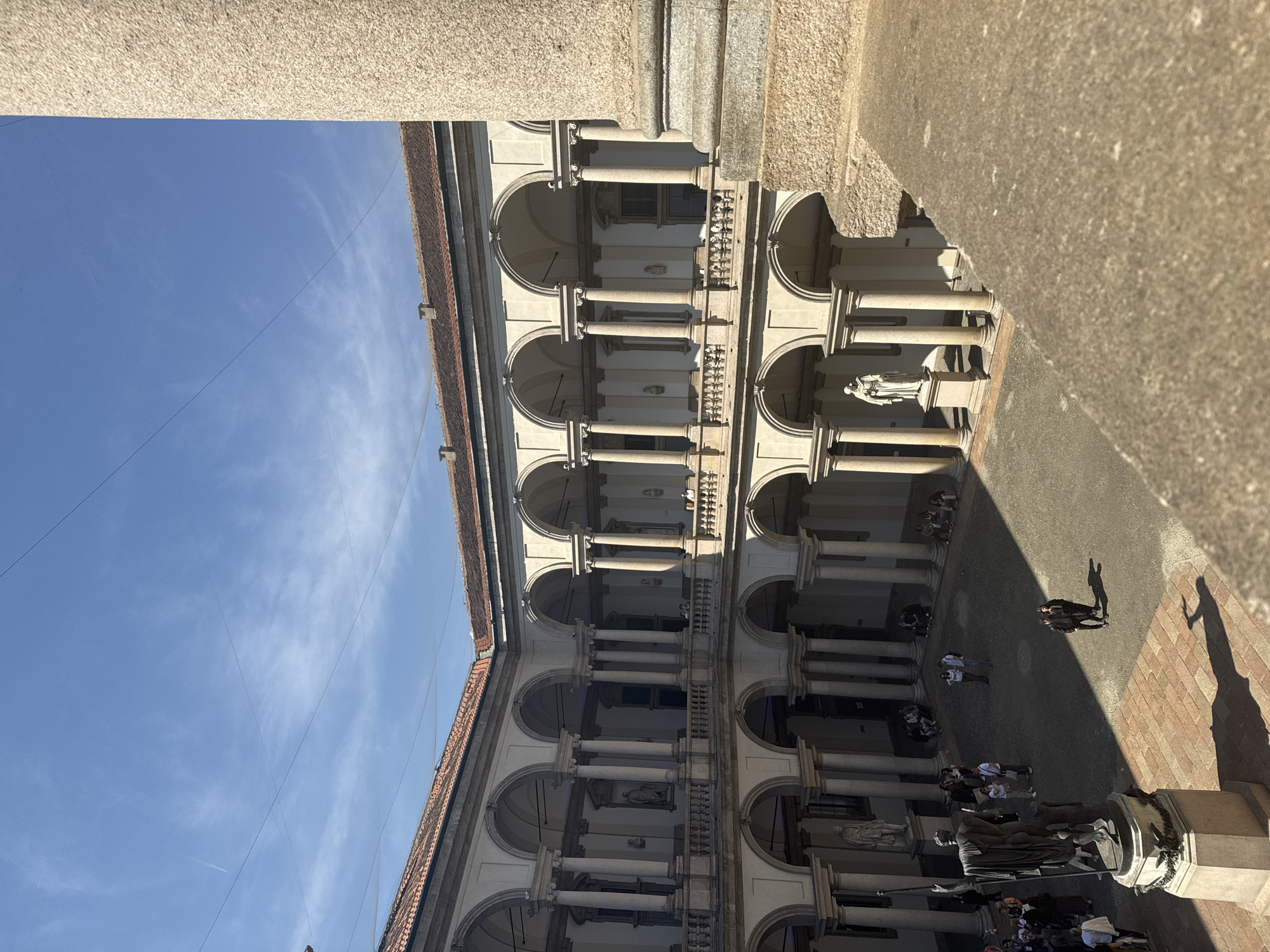 The image depicts a courtyard in Milan, Italy, likely at the Università degli Studi di Milano, a state-supported higher learning institution. The courtyard is surrounded by multi-storied buildings with arches and columns. Statues are placed within the arcades. Several individuals are seen walking or gathered in the courtyard. The sky is visible above, with a few clouds. The scene appears to be well-lit, with sunlight casting shadows in the courtyard.