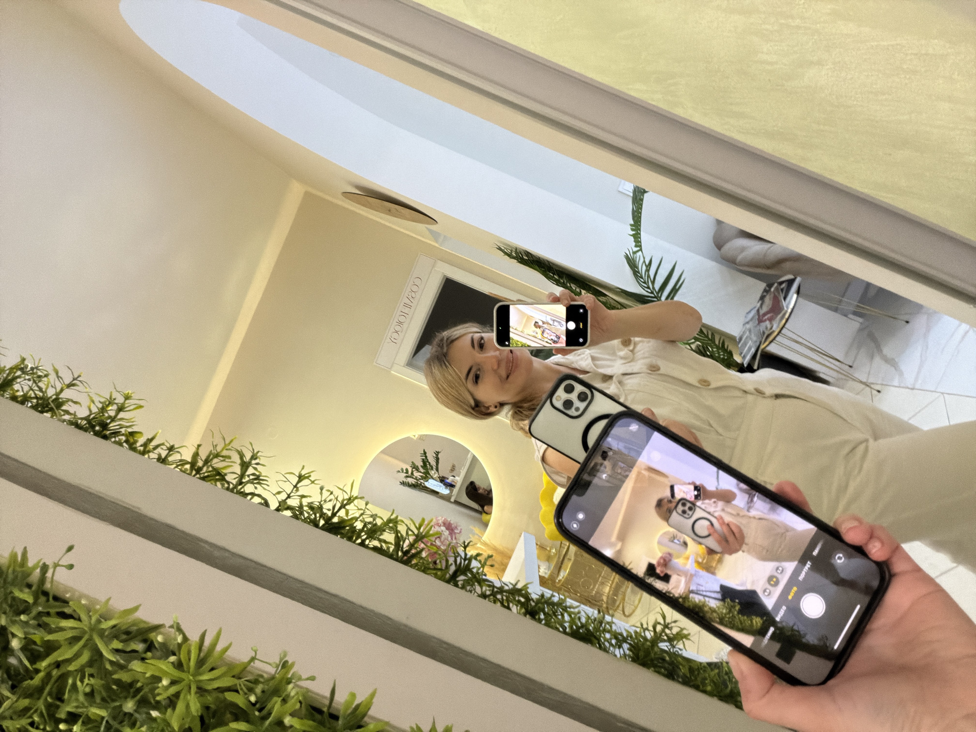 An indoor scene depicts a recursive mirror selfie. In the foreground, a hand holds a black smartphone, capturing a reflection in a large mirror. The mirror shows a blonde woman, positioned in the upper right, smiling slightly as she looks into the screen of her own white smartphone, which she is holding up to take a selfie. She is wearing a light-colored, possibly cream or beige, button-up outfit.
The reflection in the mirror reveals a bright, modern interior. On the wall behind the woman, the reversed text "COZIMONDO" is visible. A circular mirror or light fixture is also reflected in the background of the woman's selfie, showing a plant and other decorative elements. Artificial green foliage, possibly a plant wall or decorative railing, frames the bottom and left edges of the main mirror. The setting is well-lit, suggesting either bright natural light or ample artificial illumination.
Both smartphones display camera interfaces. The foreground phone's screen shows options like "PHOTO," "VIDEO," "PORTRAIT," and "PANO," with some text appearing in Cyrillic (e.g., "ФОТО", "ВИДЕО", "ПОРТРЕТ", "ПАНО"). The woman's phone screen also clearly displays her reflection within its own camera app, creating a visual loop of selfies within selfies. The entire image is tilted, giving a dynamic, angled perspective.