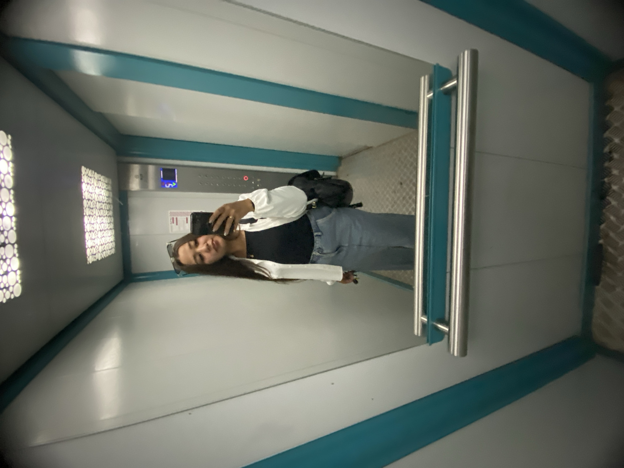 A young woman is taking a mirror selfie in an elevator. She is wearing a black tank top, a white open jacket, and a denim skirt. She has sunglasses on top of her head and is carrying a black backpack over her left shoulder. She is holding a smartphone in her right hand. The elevator has white and teal walls, a metal handrail, and a checkered floor. The ceiling has a patterned light fixture. The elevator appears to be in an indoor location, possibly a residential or commercial building. There is no information in the image to suggest it is in Ufa, Russia.