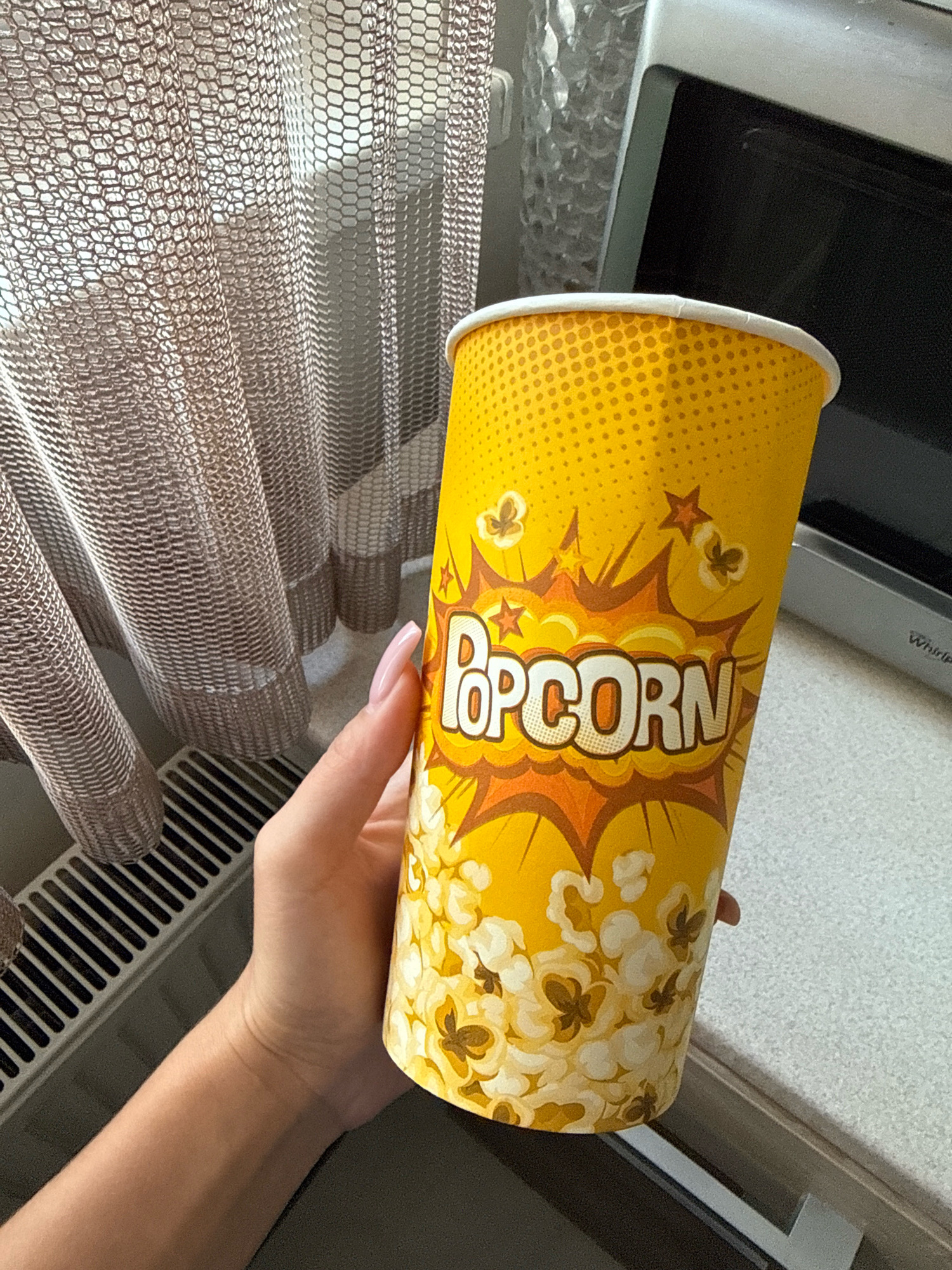 An overhead shot shows a hand holding a yellow popcorn cup. The cup is adorned with a comic book-style design featuring exploding shapes, stars, and popcorn kernels. The background includes a window with a patterned curtain, a radiator, and a microwave oven. The scene appears to be indoors, possibly in a kitchen or living area. The image does not provide any information that would allow for its location in Chișinău, Moldova, to be determined.