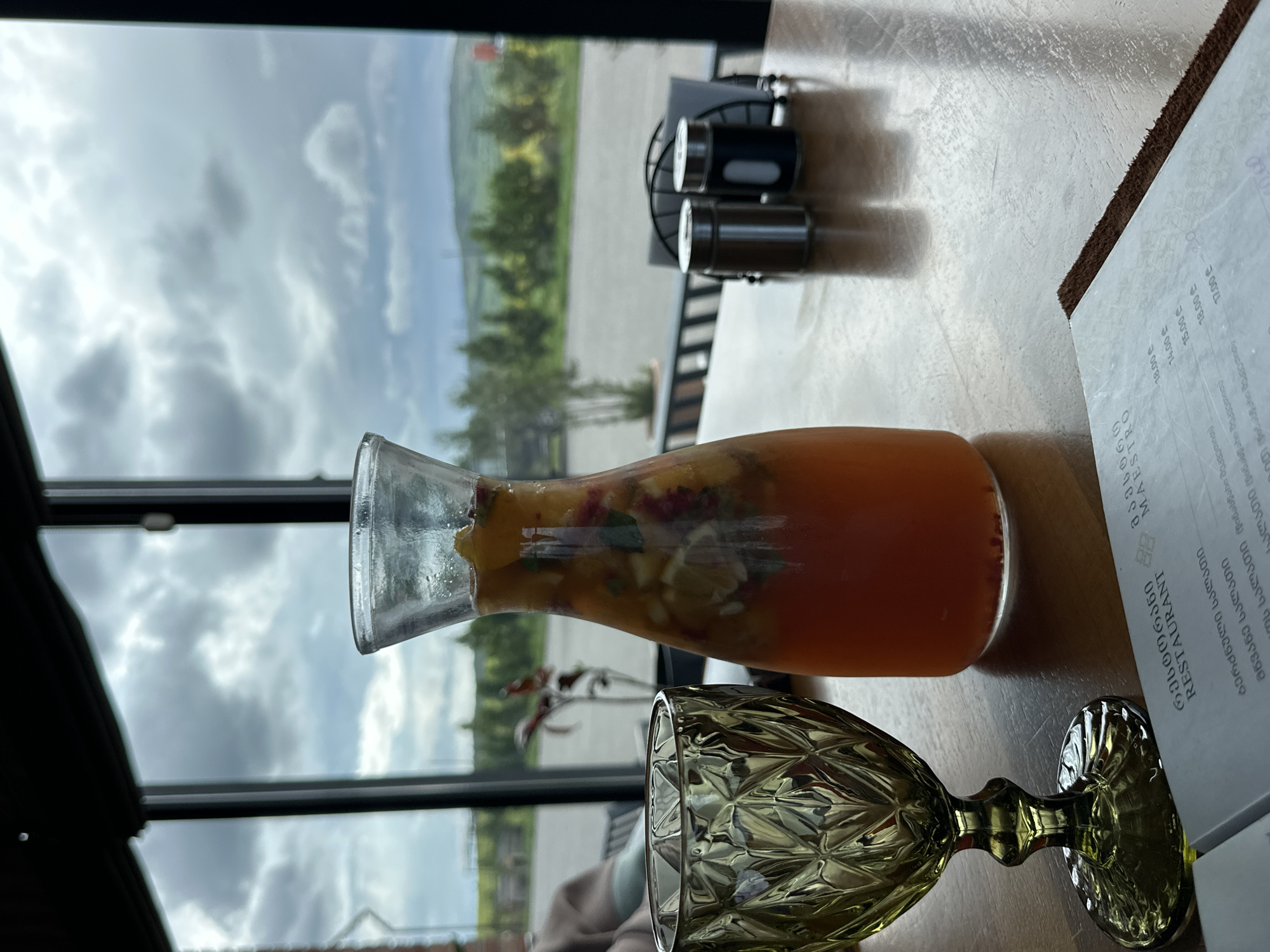 A close-up, eye-level, indoor shot shows a glass decanter filled with a reddish-orange beverage containing sliced fruit, along with a stemmed glass, a restaurant menu, and salt and pepper shakers on a table in a restaurant.
The table is light-colored and appears to be made of wood or laminate. The glass decanter is mostly filled with a drink. The stemmed glass is ornate and empty. The menu is partially visible in the upper-right corner and features text in Georgian (restaurant MAESTRO). The shakers sit toward the back edge of the table.
The background is an exterior view of clouds, sky, trees, and grass, seen through the restaurant's window. The location is Marneuli, Georgia.