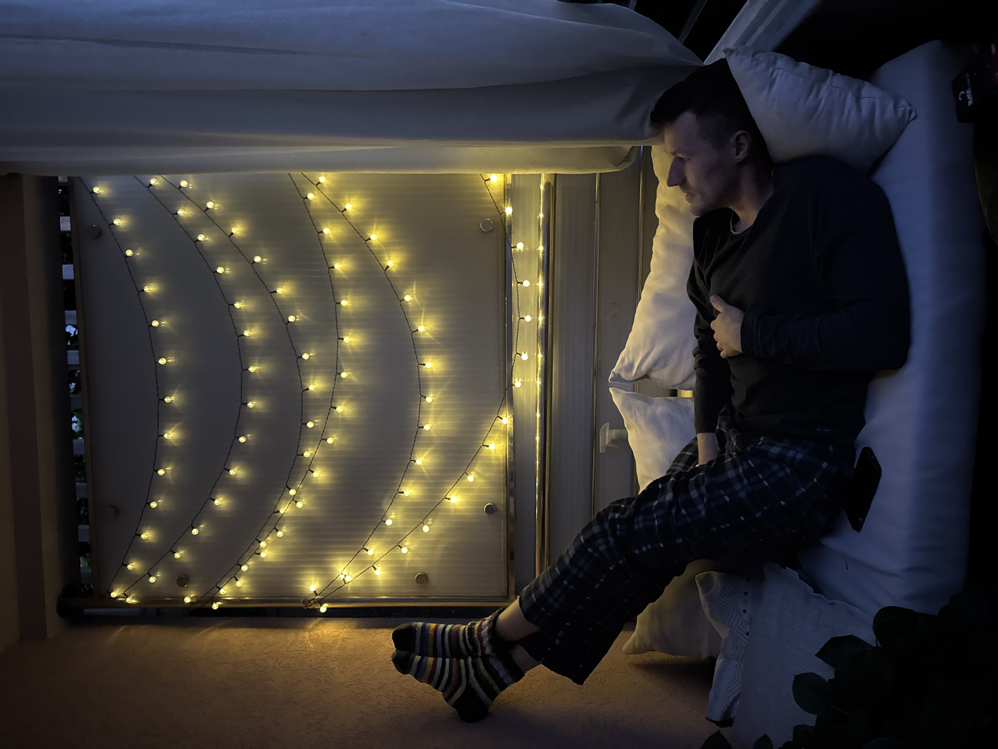 A person is lying on a makeshift bed inside a balcony enclosed with blinds and decorated with string lights. The person is wearing a dark long-sleeved shirt and plaid pajama pants, and striped socks. They are lying on white pillows with their hand resting on their chest. The room is dimly lit by the string lights. The location appears to be a balcony setting, potentially in a residential building in Budva, Montenegro, considering the presence of blinds and balcony railing. The image depicts a scene of rest or relaxation in a confined space.