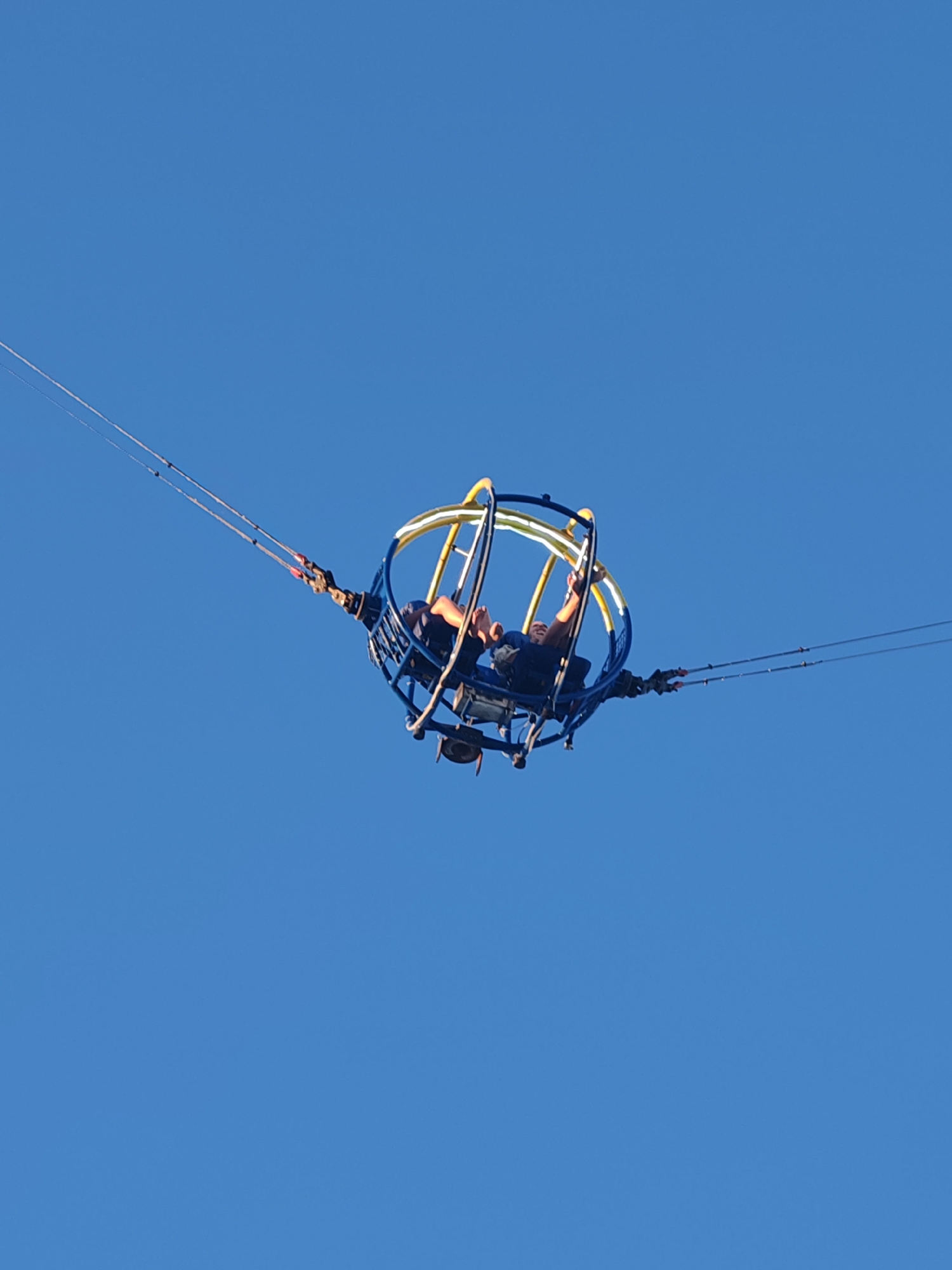 Two people are seated in a "slingshot" amusement ride at Nesebar, Bulgaria. The ride is elevated against a clear blue sky. The circular, caged structure is secured by cables extending to the left and right. The structure has yellow and blue accents. The riders are facing upwards.