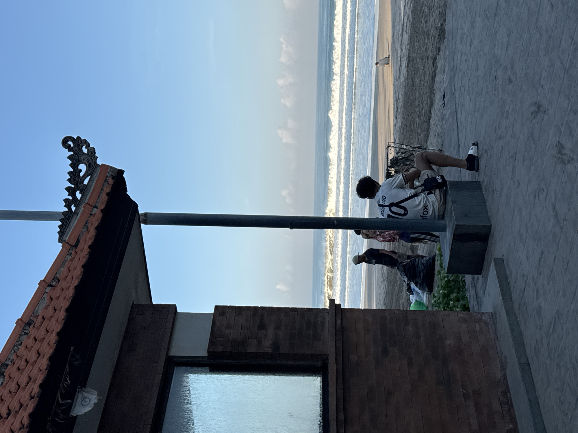 The image captures a peaceful coastal scene in Kuta, Indonesia, bathed in the soft light of what appears to be late afternoon or early evening under a clear blue sky.
On the left, a traditional Balinese-style building is partially visible. It features a terracotta tiled roof with an intricately carved dark brown or black decorative element adorning its peak. The lower part of the building shows a wall clad in brick-like material and a large window with textured glass. A small white flag or cloth with a circular motif hangs from the building's side.
A dark metal pole stands centrally, leading towards the beach. A man is seated on a low stone wall or bench, his back mostly to the camera, facing the ocean. He wears a white and black jersey displaying the number "10", and text that reads "RAFA RAO" and "fox". He is dressed in shorts and sandals, appearing relaxed as he looks towards the sea. Near him, some green foliage adds a touch of nature. Behind him, closer to the shore, other indistinct figures are visible, including one person in a plaid shirt.
To the right, the expansive ocean stretches out, with white-crested waves rolling onto a sandy beach. In the distance, a solitary figure in light clothing stands by the water's edge. The clear sky with a few wispy clouds enhances the serene and leisurely atmosphere of the scene.