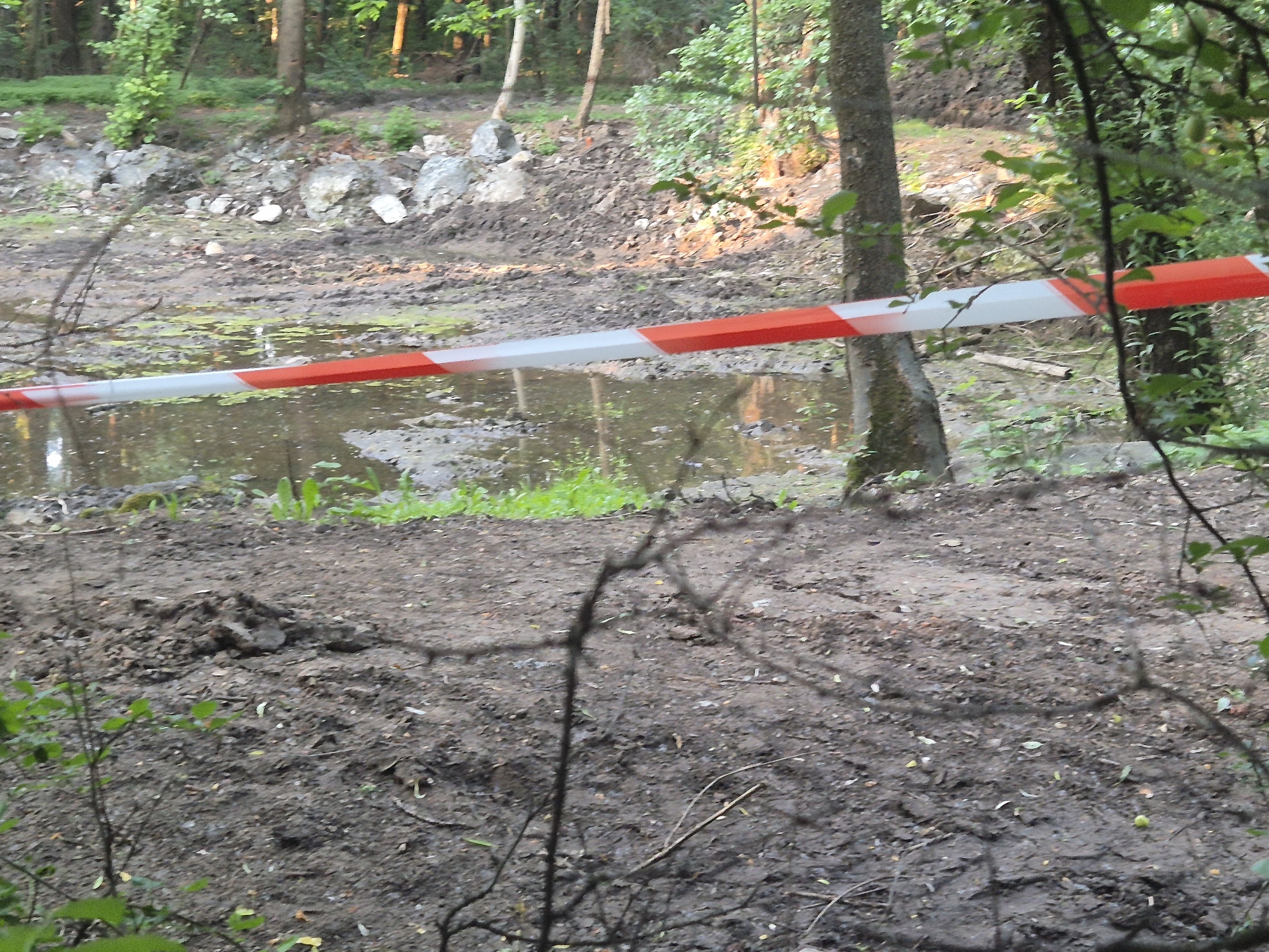 The image depicts a muddy area within a forest, likely a park or a construction site, showing signs of recent excavation or disturbance. A section of red and white striped caution tape is visible in the foreground, stretched horizontally across the frame. The tape is positioned in front of a murky pool of water and a pile of dirt. In the background, trees and foliage are abundant, suggesting a natural or semi-natural environment. The presence of caution tape implies that the area is considered potentially hazardous or restricted. The ground is uneven and covered with mud, rocks, and debris, indicating that work may have been done or is planned for the area. The lighting suggests it might be daytime, possibly overcast or late afternoon given the shadows. The scene is located in a forested area within Sofia, Bulgaria.