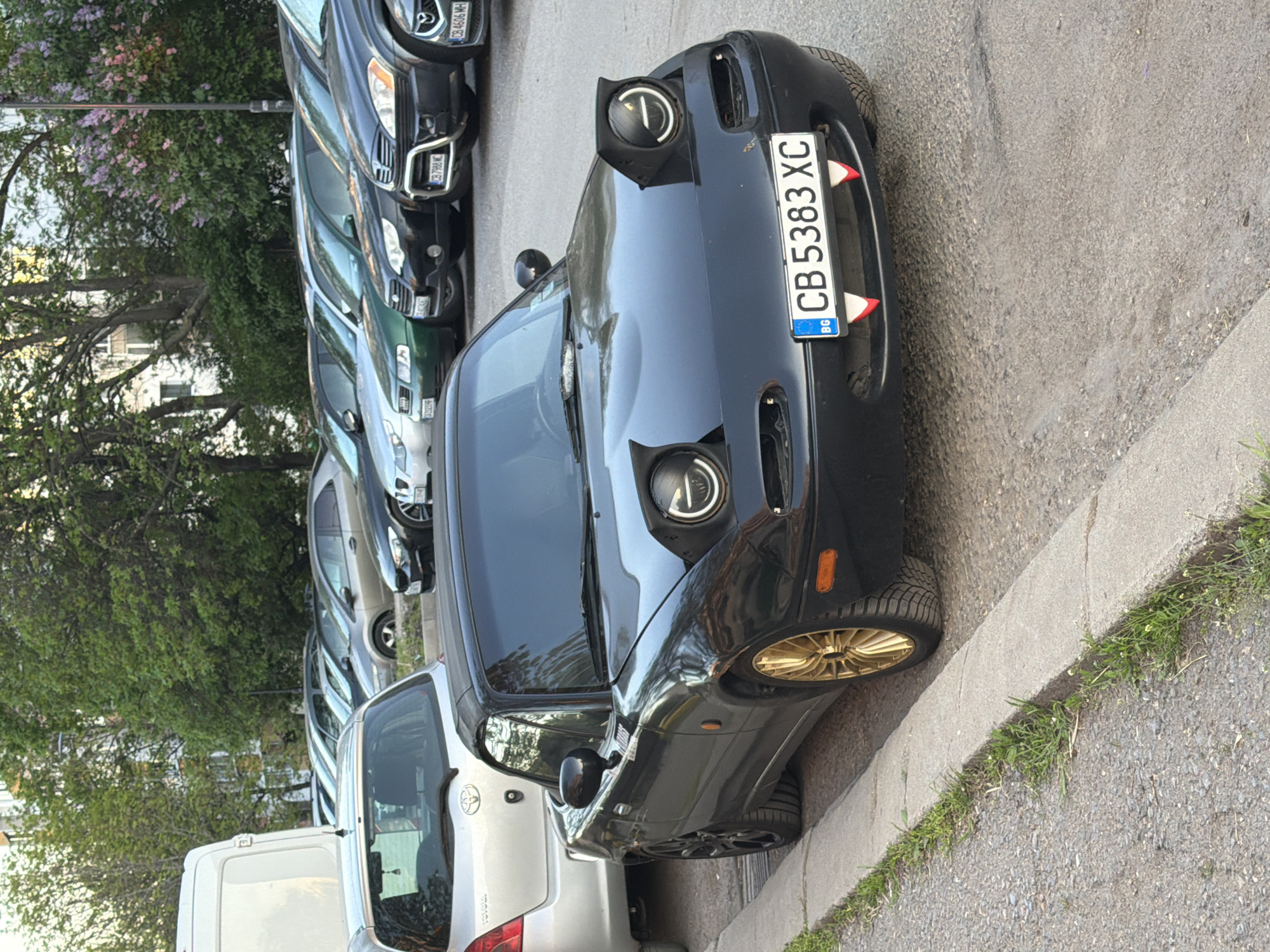 The image shows a black Mazda Miata NA parked on a street in Sofia, Bulgaria. The car has gold-colored wheels and a Bulgarian license plate that reads "CB 5383 XC" with two white and red upside-down triangles attached to the plate. The car is parked next to a silver Toyota Yaris and a row of other parked cars, including a green SUV and several dark-colored sedans. A concrete curb separates the parking space from a patch of grass and the street. Trees with blooming purple flowers are visible in the background. The scene appears to be a typical urban parking situation.